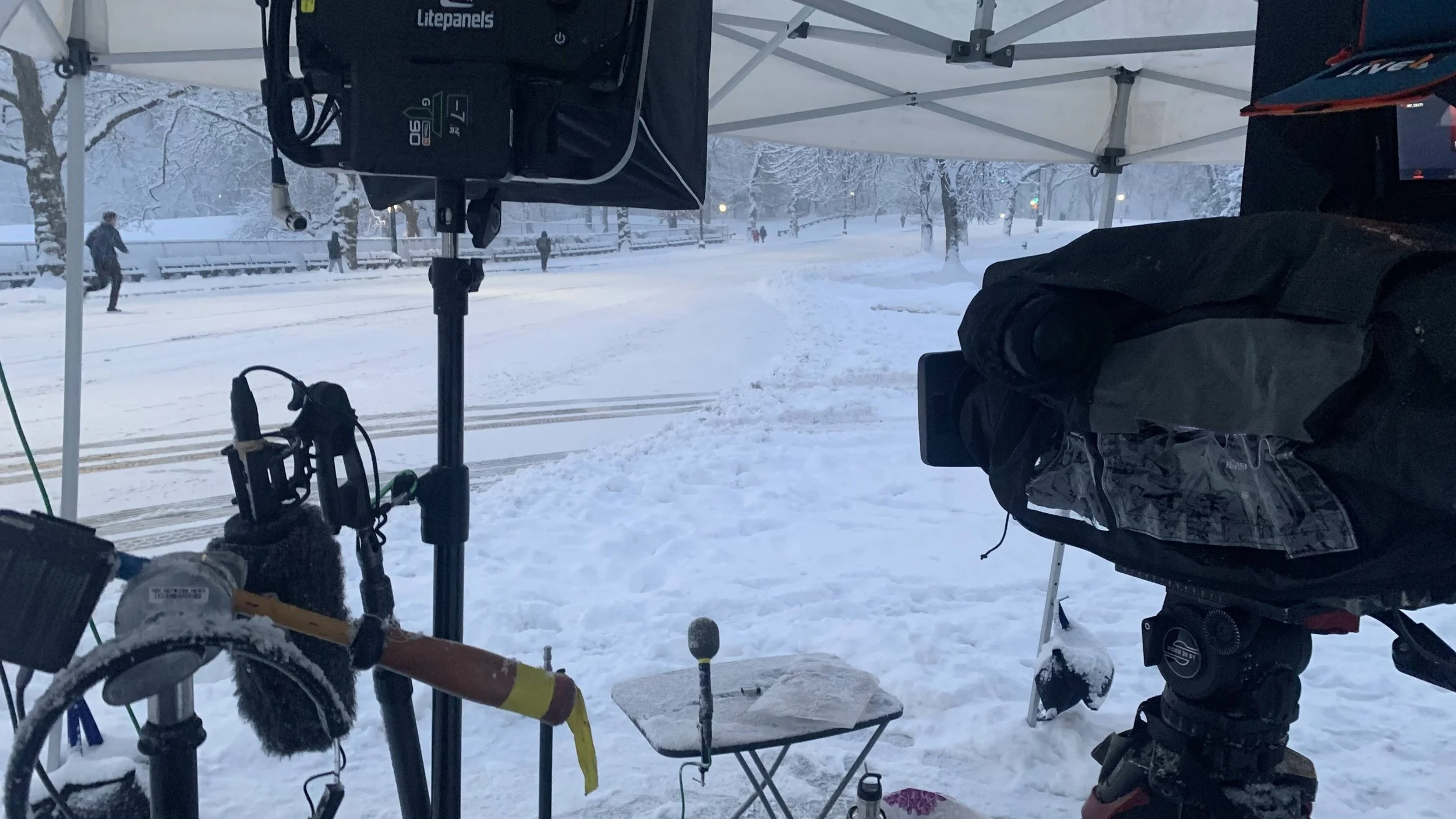 A film crew setup under a canopy in a snowy park, with camera equipment, a small table, and snow-covered trees and paths in the background.