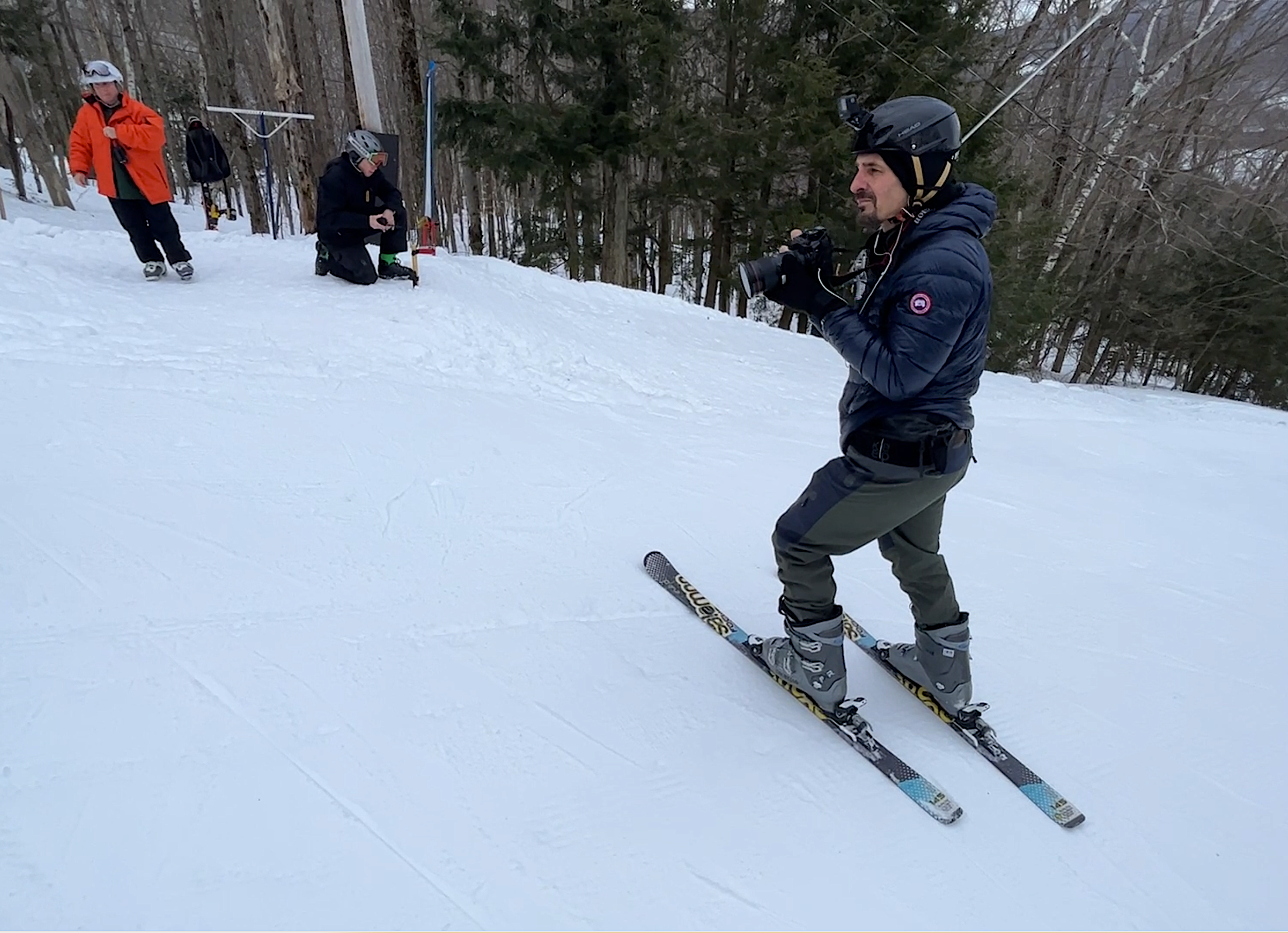 A man in a black helmet and blue winter jacket skiing on snow, with two people in the background near trees, one standing and one kneeling.