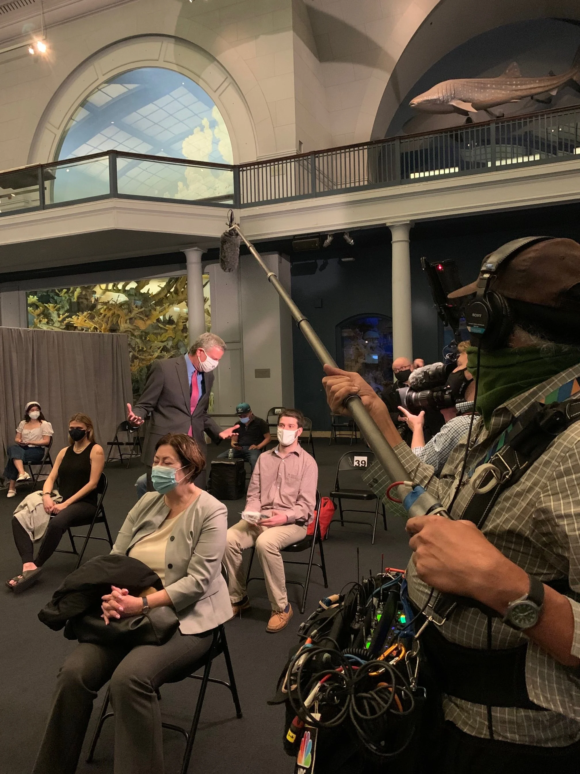 People sitting and standing in a room with high ceilings, some wearing face masks, as a person with a camera films the scene.