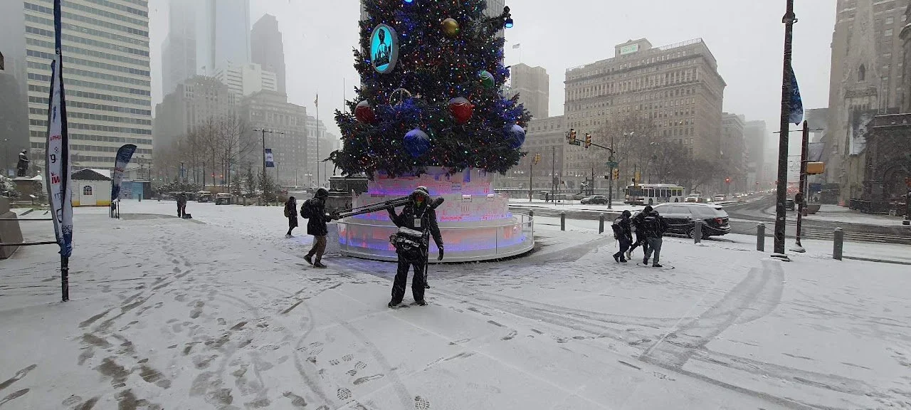 A snowy city square with a large decorated Christmas tree at the center. Several people are walking around, some carrying skis, with buildings and traffic in the background. It appears to be during winter with overcast skies.