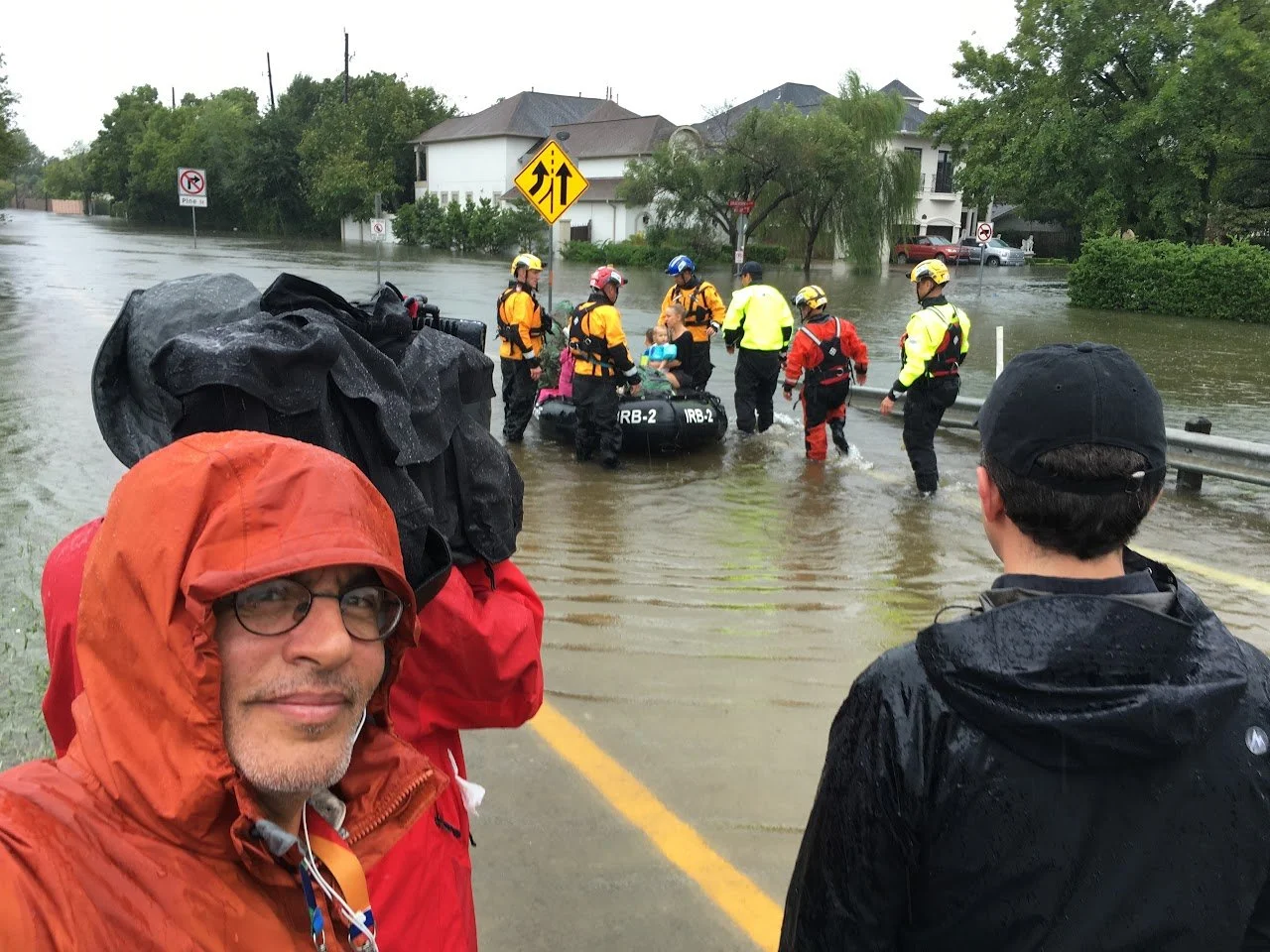 Rescue team wading through floodwaters to assist people in a partially submerged neighborhood, with houses and trees in the background.