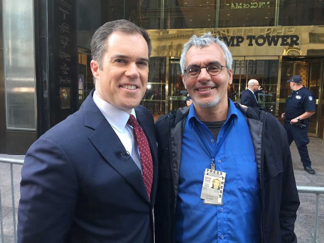 Two men smiling outside Trump Tower in New York City. One in a suit and tie, the other with a press pass around his neck, in front of a glass entrance with people and police officers in the background.