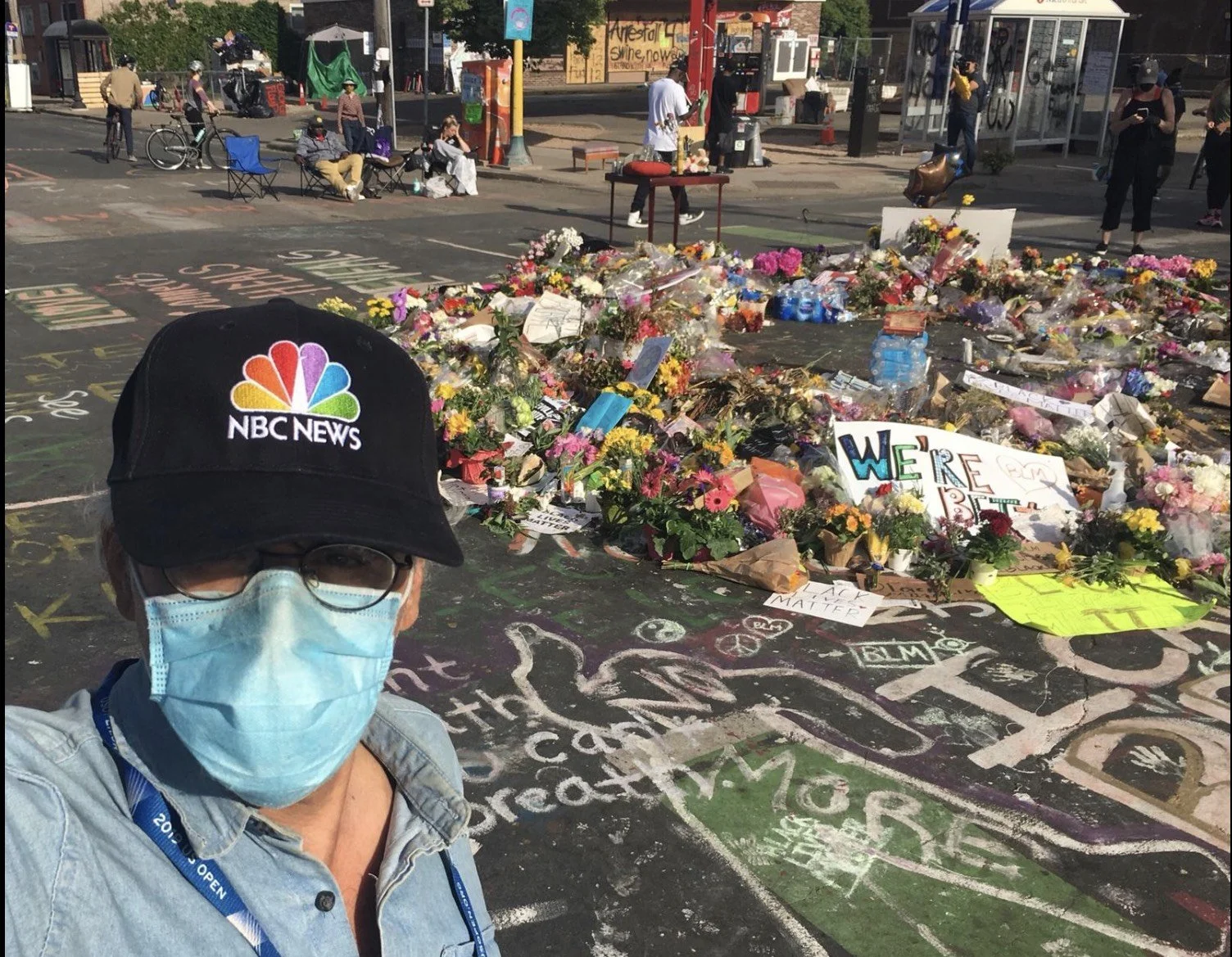 Person wearing a NBC news cap, glasses, face mask, and denim shirt taking selfie at a memorial site with flowers and signs, in a city street with people and bicycles in the background.
