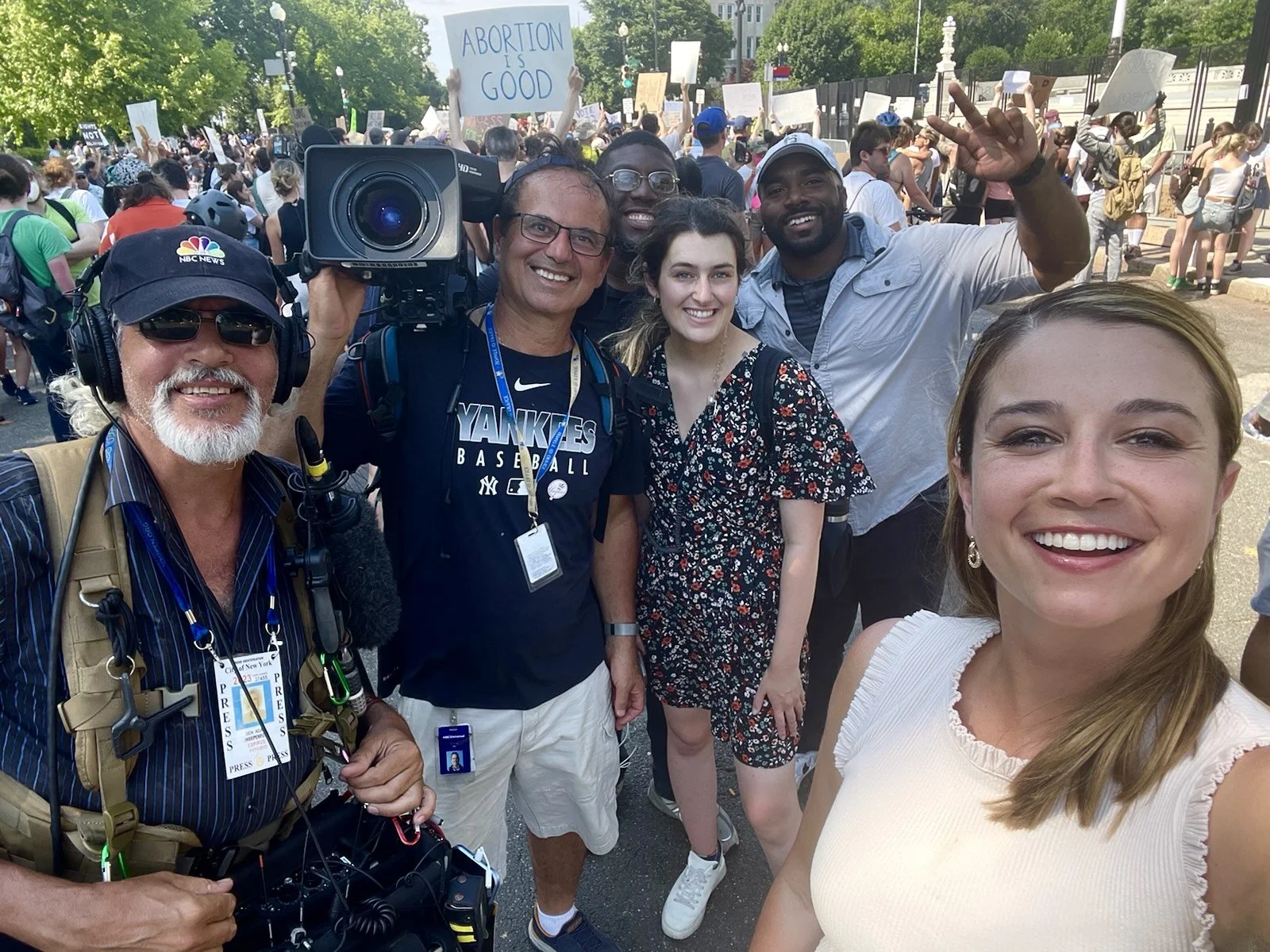 A group of diverse people smiling at a protest or rally, with a sign reading 'Abortion is Good' in the background.