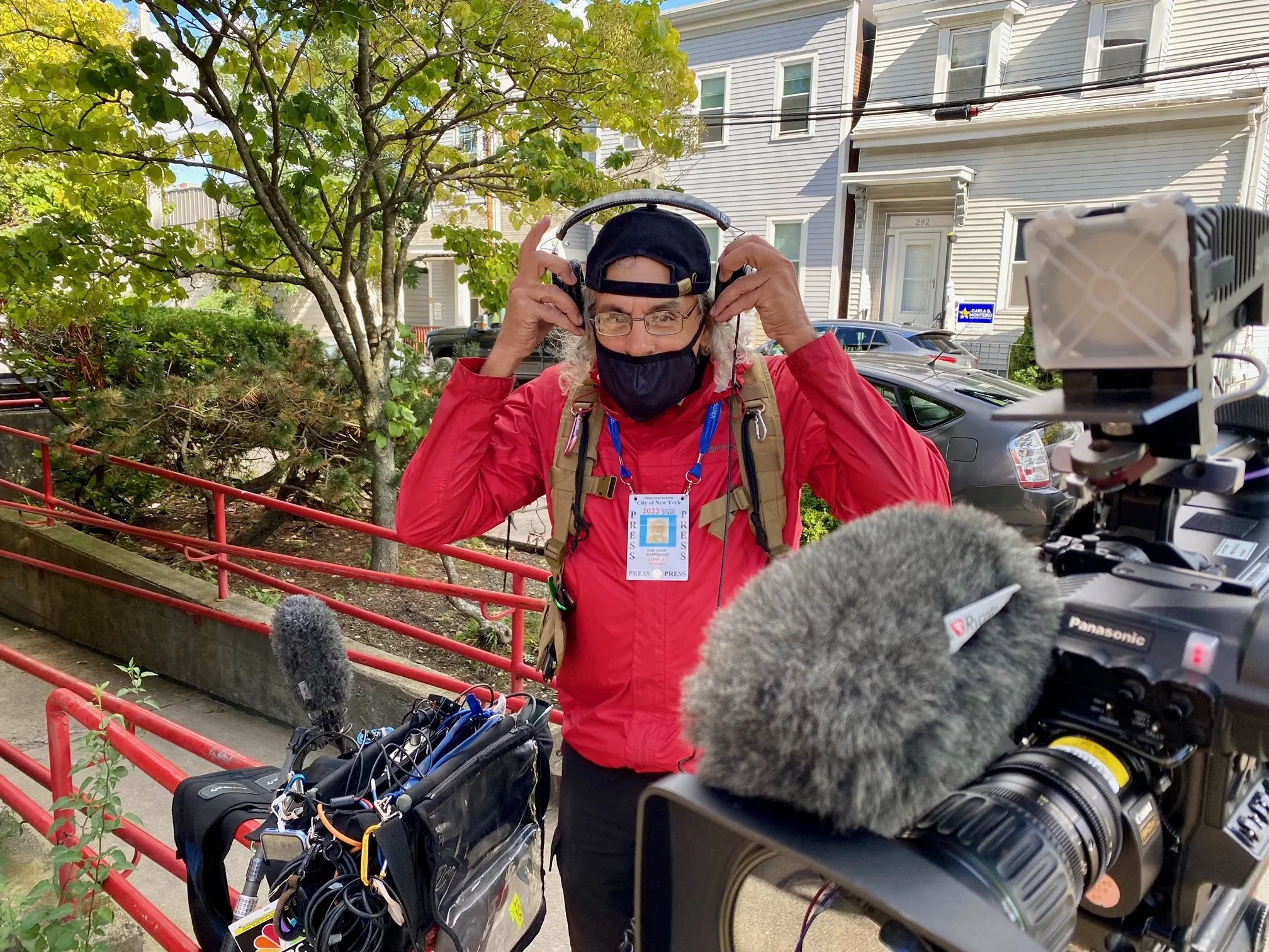 A person wearing a hat, glasses, and a face mask adjusts large headphones while standing outdoors among trees and residential buildings, with a professional video camera and audio equipment nearby.