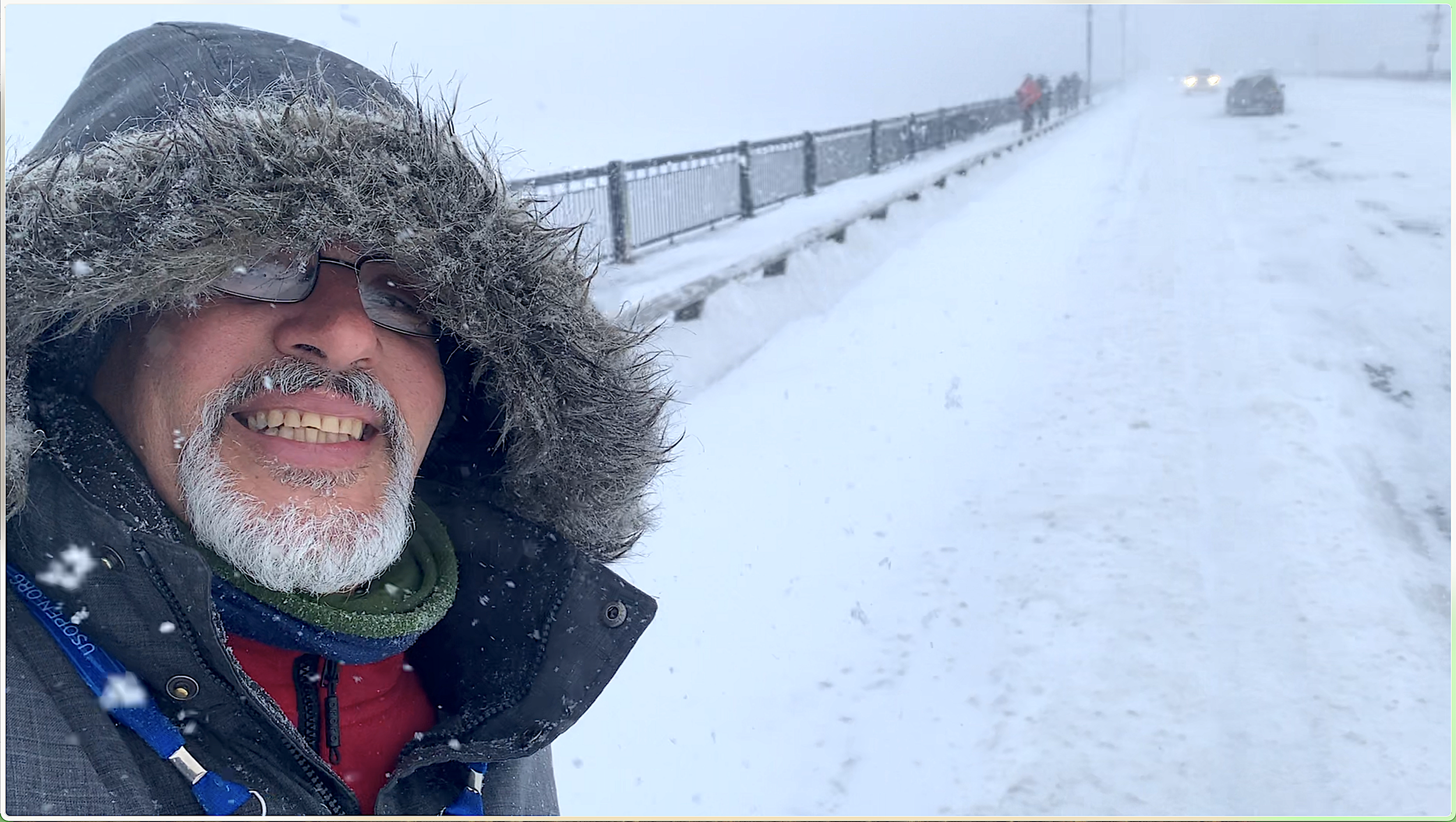 A man with a white beard and glasses taking a selfie in a snowy landscape, wearing a hoodie with a fur-lined hood and a winter coat, with snow falling around him.