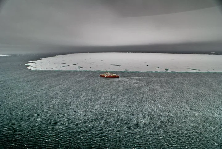 Cargo ship navigating icy waters near a large ice formation under a cloudy sky.