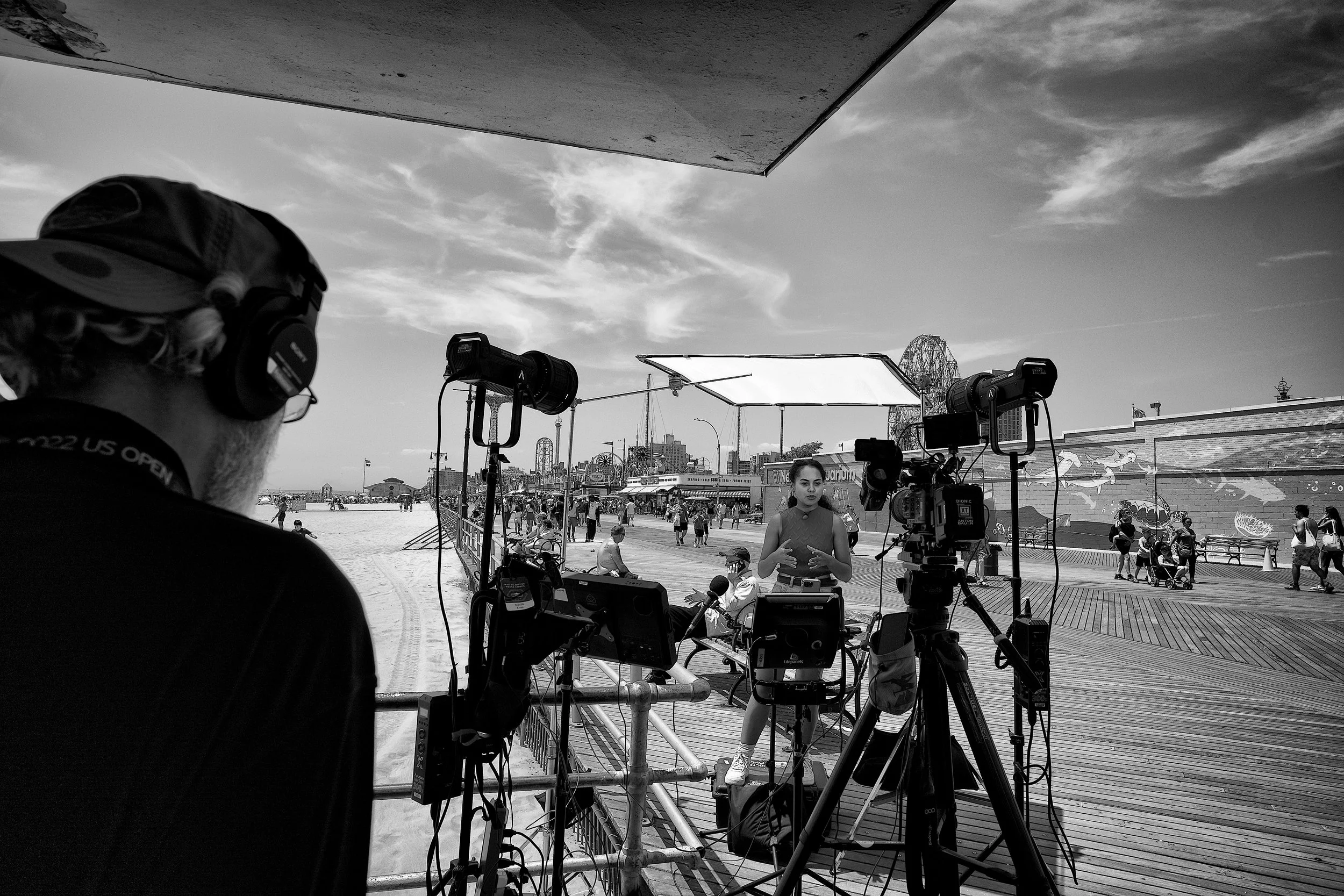 A woman is being recorded by a film crew on an outdoor boardwalk at an amusement park with roller coasters in the background, and people walking around.
