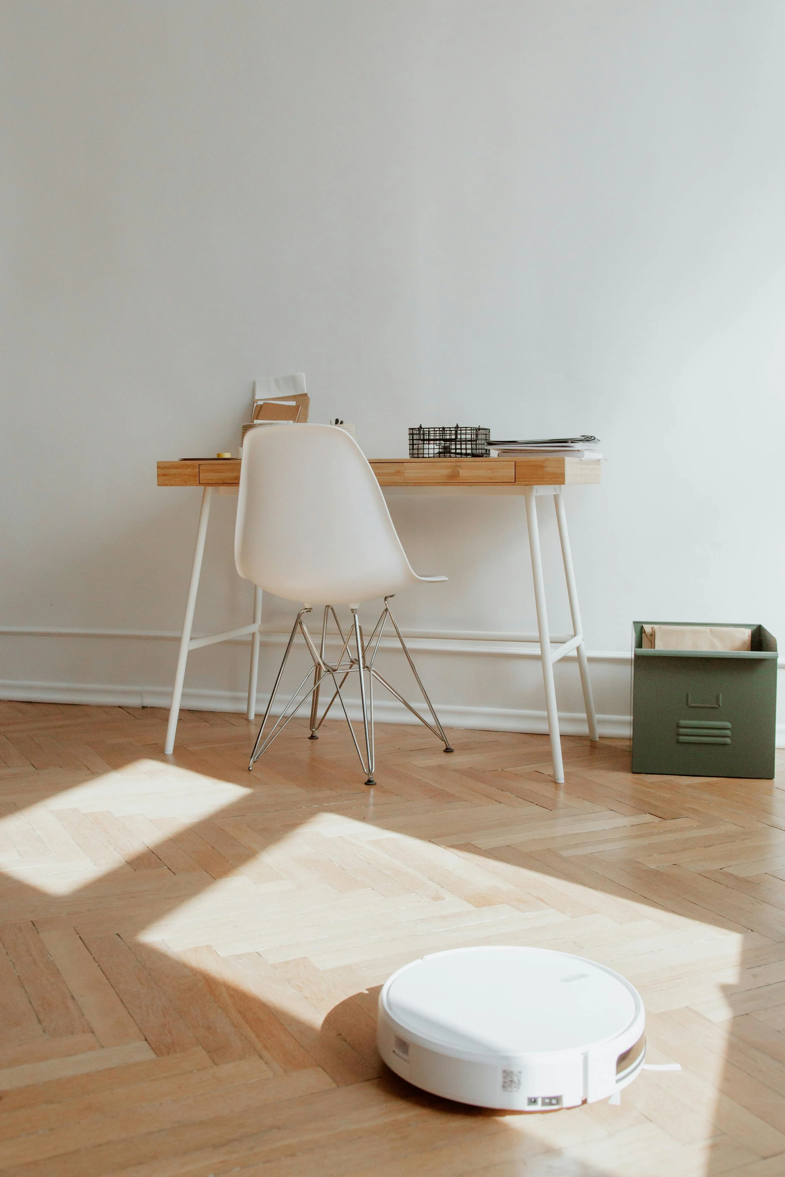 White roomba running in an office with sunlight shining in, a wood desk, white chair and green filing cabinet holding paperwork.