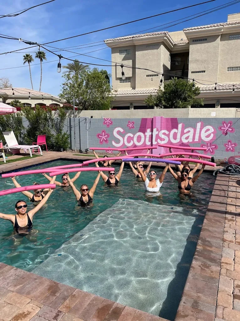 Group of women in a pool holding pink pool noodles overhead, with a Scottsdale sign painted on a wall in the background for Aqua Yoga and Barre session.