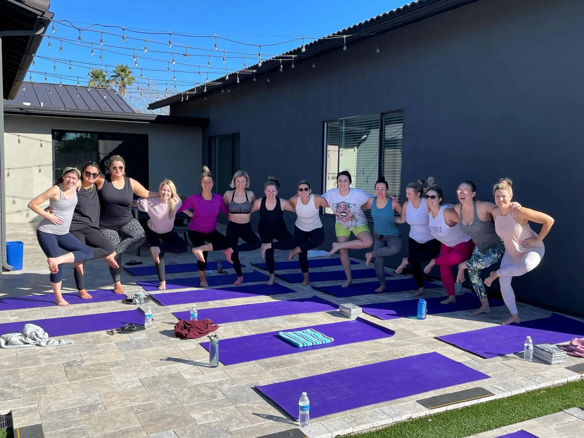 A group of women practicing yoga outdoors on purple mats, standing in a line with their arms around each other, on a patio with string lights overhead and a modern house in the background.