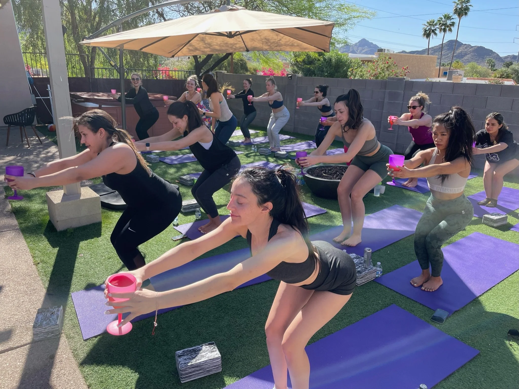 A group of  bachelorettes doing outdoor yoga or fitness class, holding pink and purple glasses, with some holding water bottles, on purple mats under a large umbrella in sunny weather, in a backyard practicing wine and yoga.