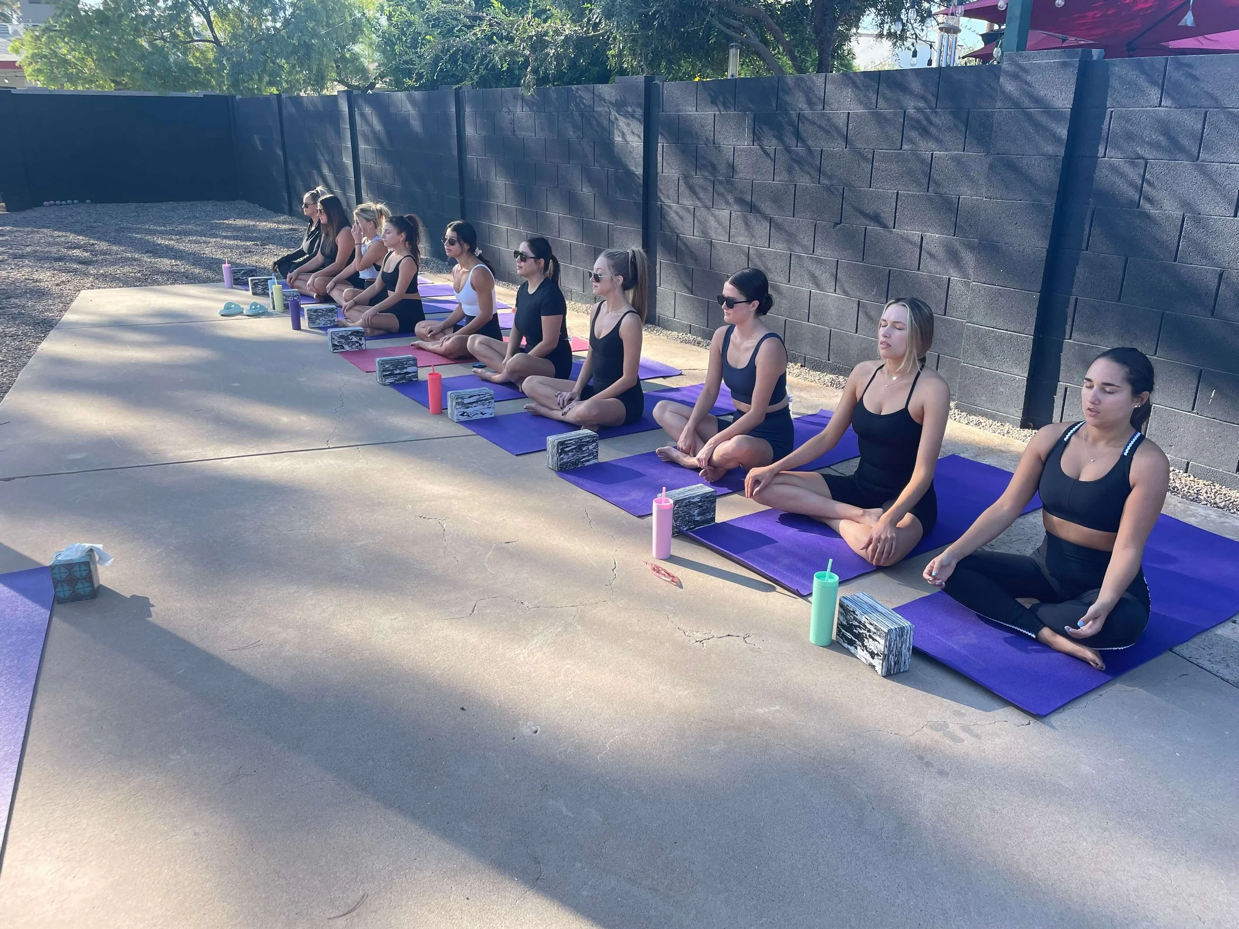 A large group of women sitting on purple mats meditating with matching black outfits and colorful water bottles and the bride in white.