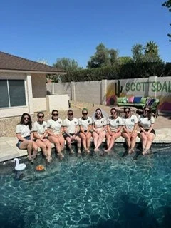 A group of friends sitting in a pool's edge wearing matching tshirts that celebrate the bride for a water yoga celebration