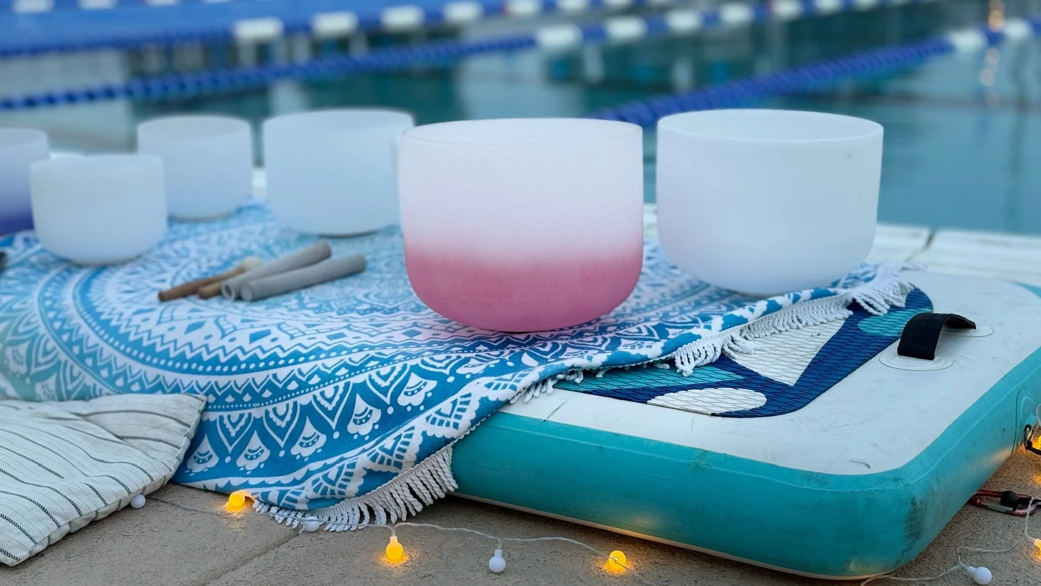 A set of white and pink crystal singing bowls on a blue and white patterned cloth next to a swimming pool, with string lights on the ground for a private Floating Sound Bath.