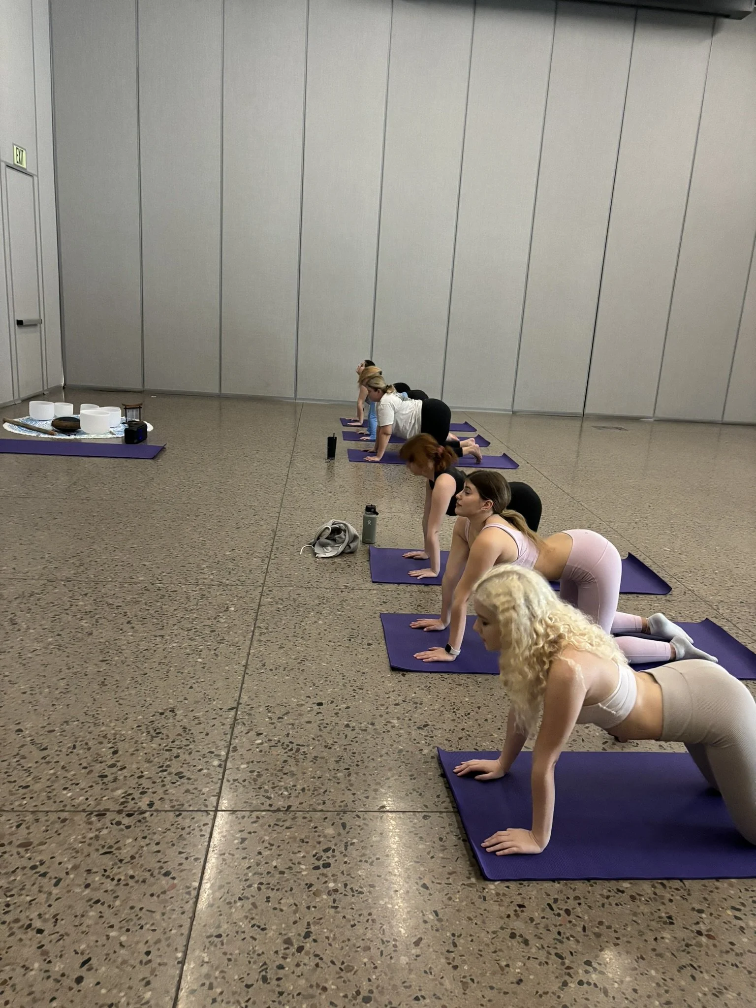Five women practicing yoga on purple mats in a spacious corporate settings indoor room with gray walls, in a table position.