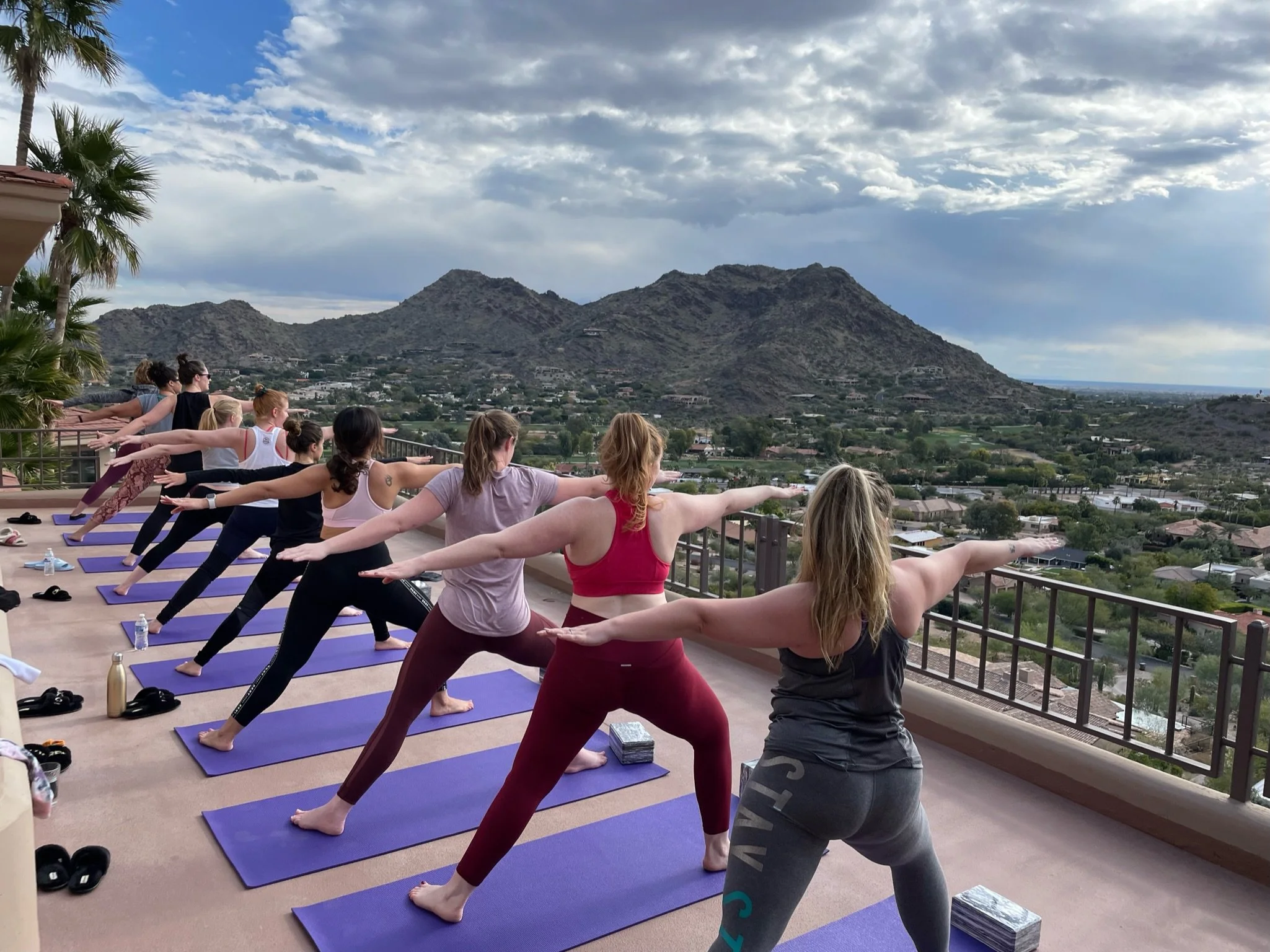 Group of women practicing a private bachelorette yoga session on purple mats on a balcony with mountain view.