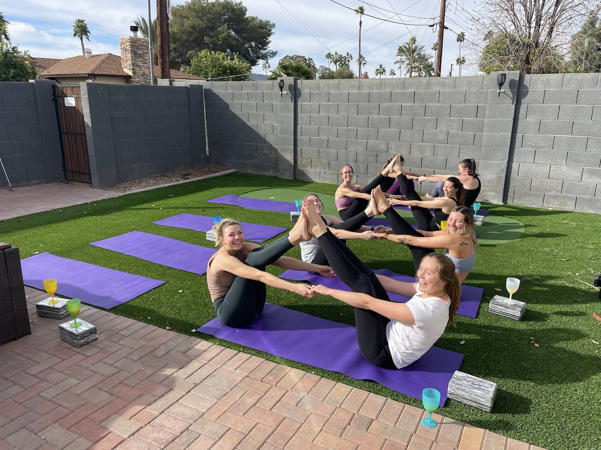 A group of six women practicing partner yoga outdoors on purple mats in a backyard enclosed with gray concrete block walls. They are sitting facing each other with legs extended and holding each other's feet, smiling and engaging in a partner stretch