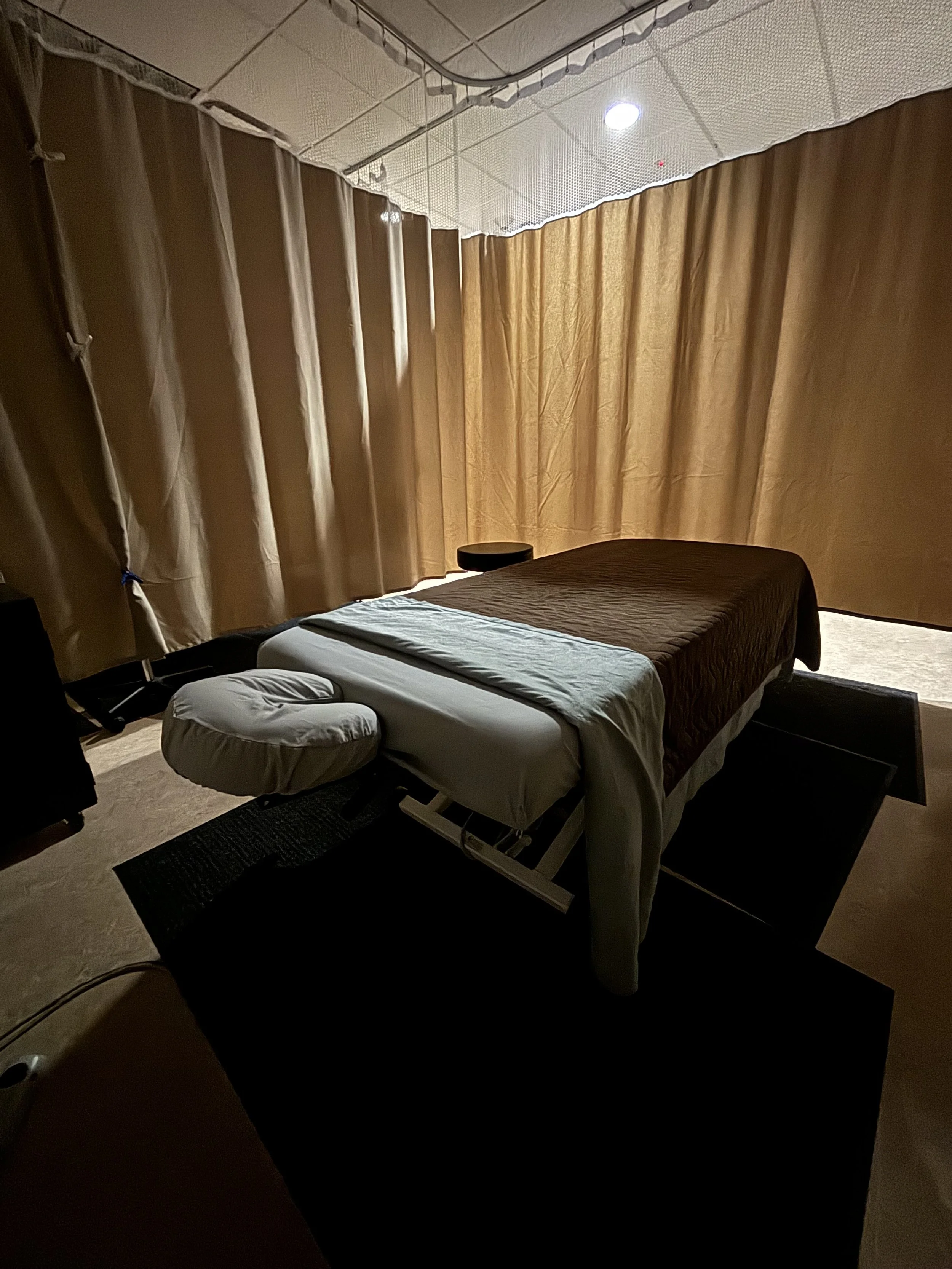 Massage table with face cradle in a dimly lit massage room, beige curtains, and a small round stool.