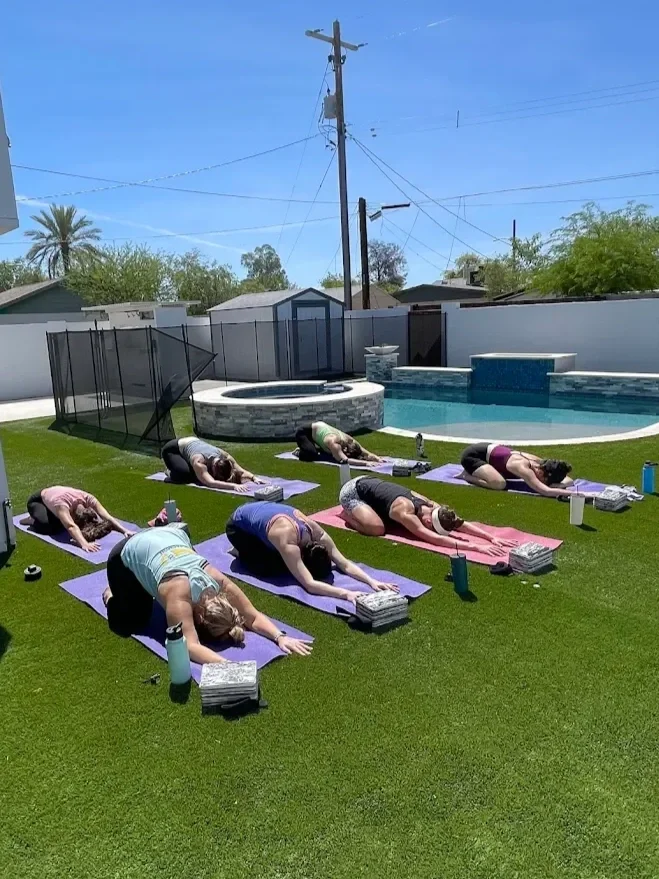 Group of people in child pose yoga positions on mats, outdoors by a pool on a sunny day.