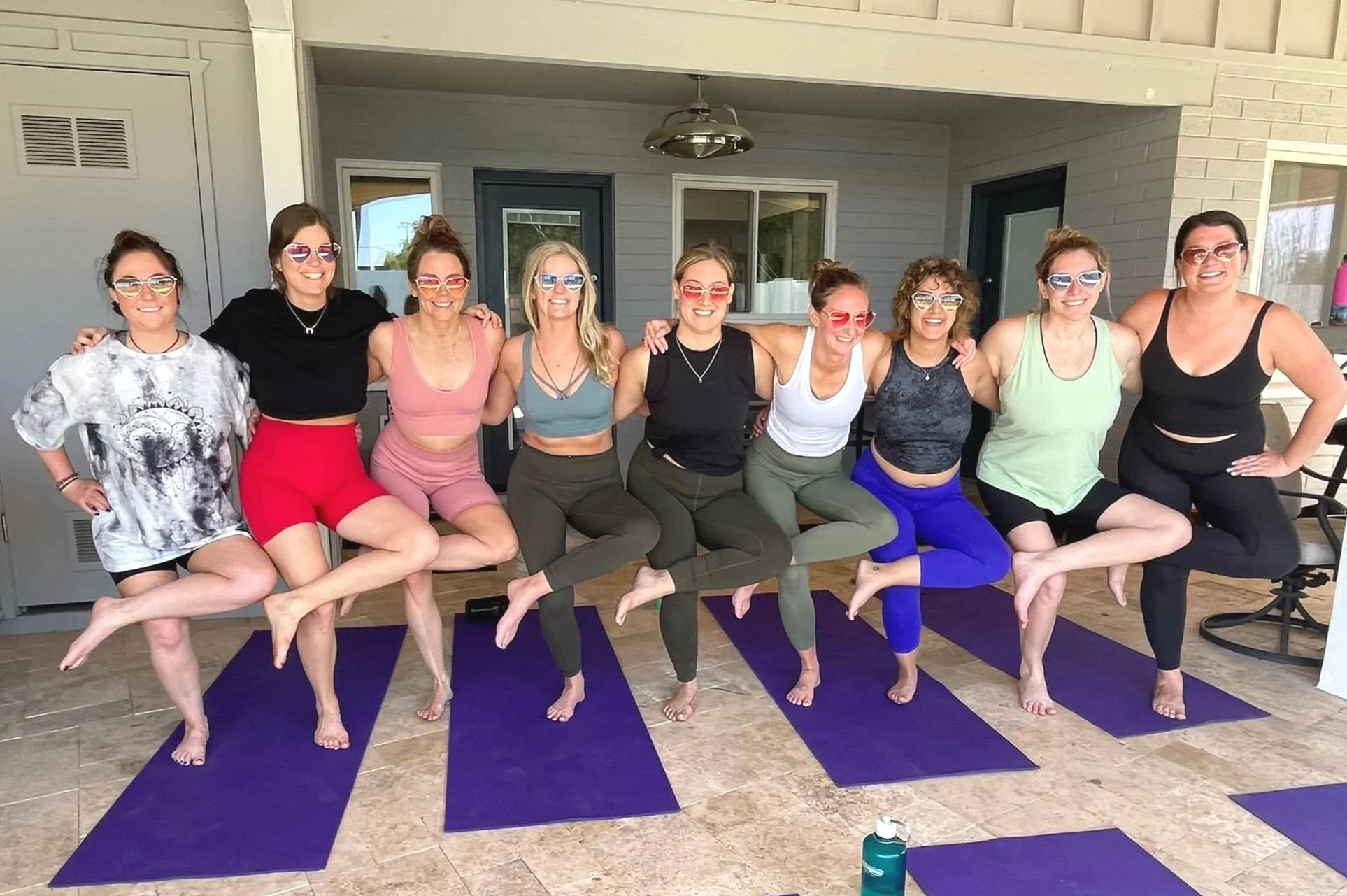 A group of ten women in a figure four standing on yoga mats, smiling, with arms around each other, all wearing sunglasses, on a patio with a modern house in the background.