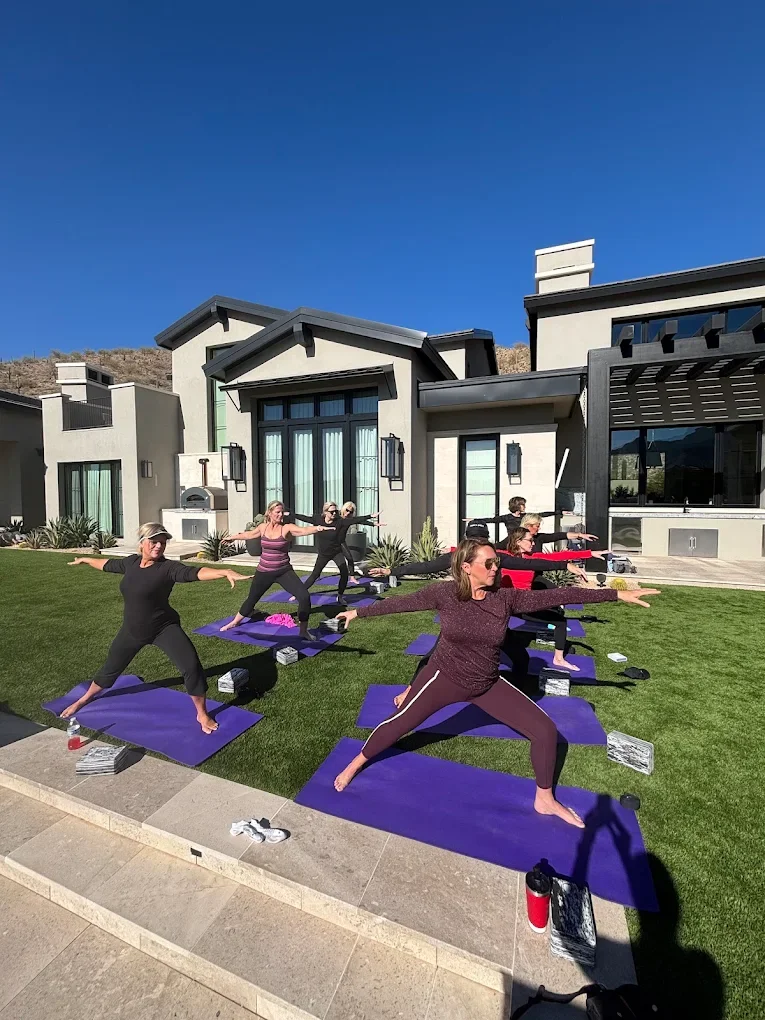 A group of women participating in outdoor yoga class on a grassy lawn in front of a modern house, practicing yoga poses on purple mats celebrating a family reunion.