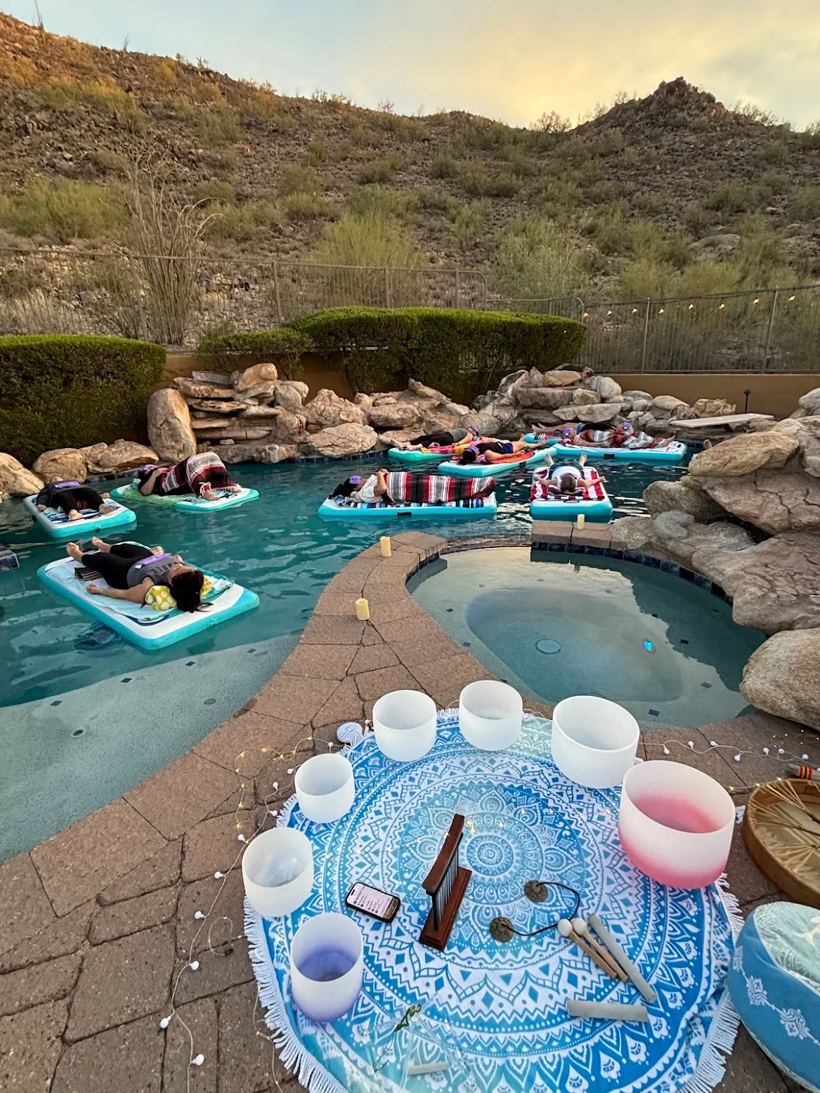 A group of people lying on floating mats in a backyard pool, possibly during a meditation or relaxation session, with a scenic mountain backdrop, and a decorative setup with singing bowls on a blue patterned cloth nearby.