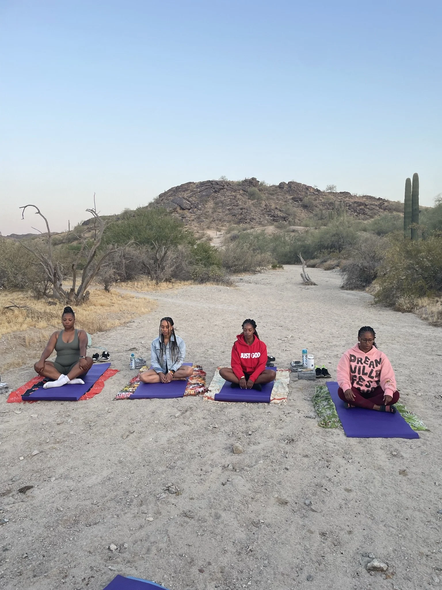 Four women practicing yoga outdoors on yoga mats in a desert landscape with mountains, cacti, and sparse trees in the background.