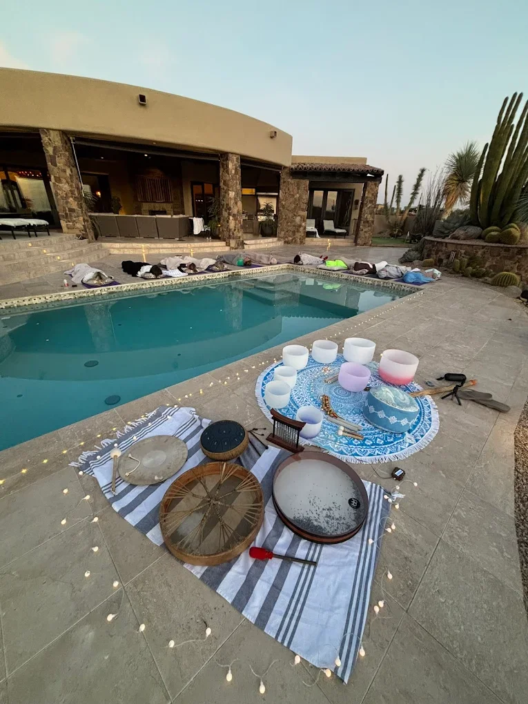 Outdoor house with a swimming pool, people lying on the steps, and a musical setup with bowls, a finger cymbal, and a singing bowl on a striped towel surrounded by string lights.