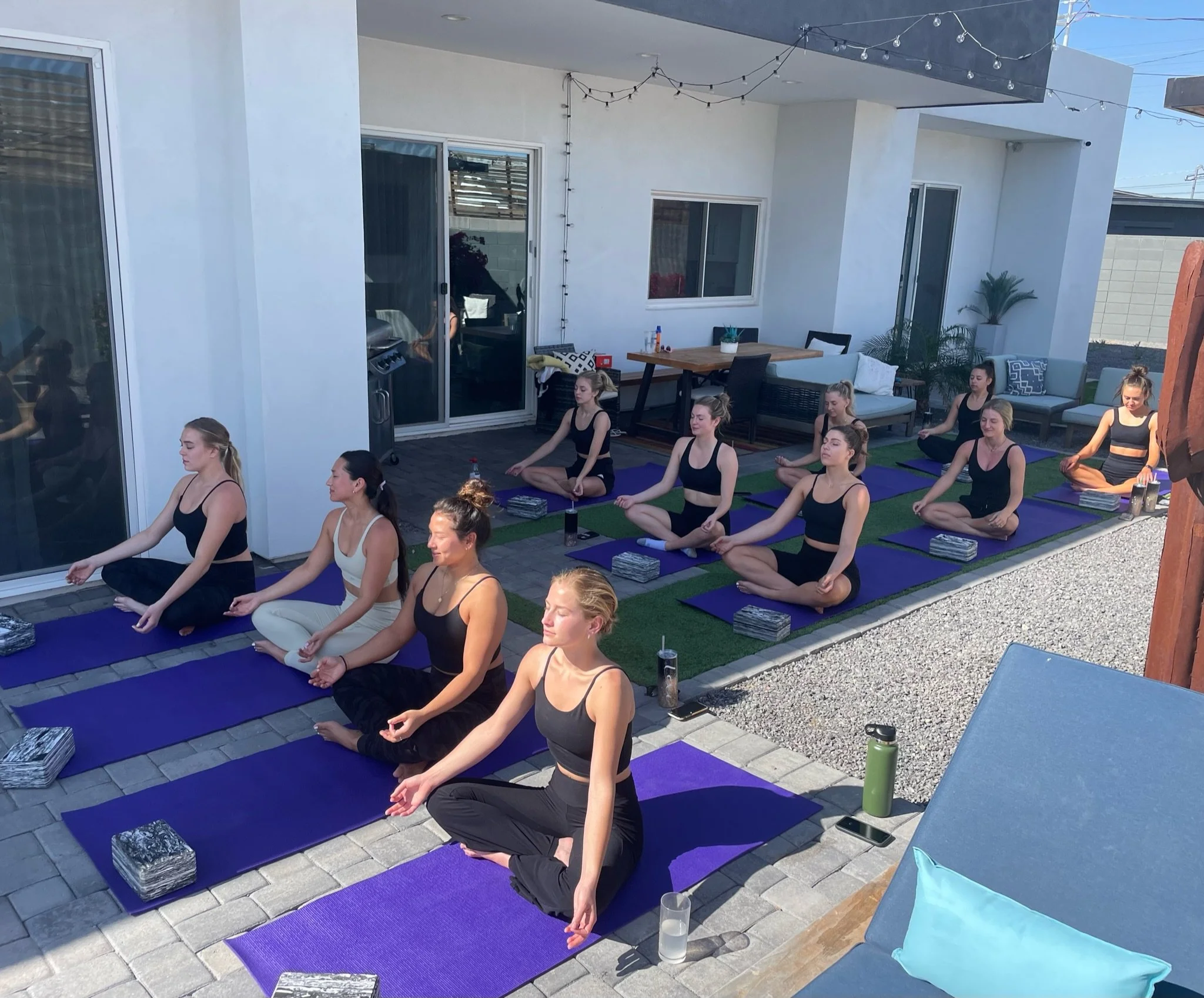 A group of women participating in an outdoor yoga class on a patio, seated on purple mats in a cross-legged position with eyes closed, practicing meditation or mindfulness.