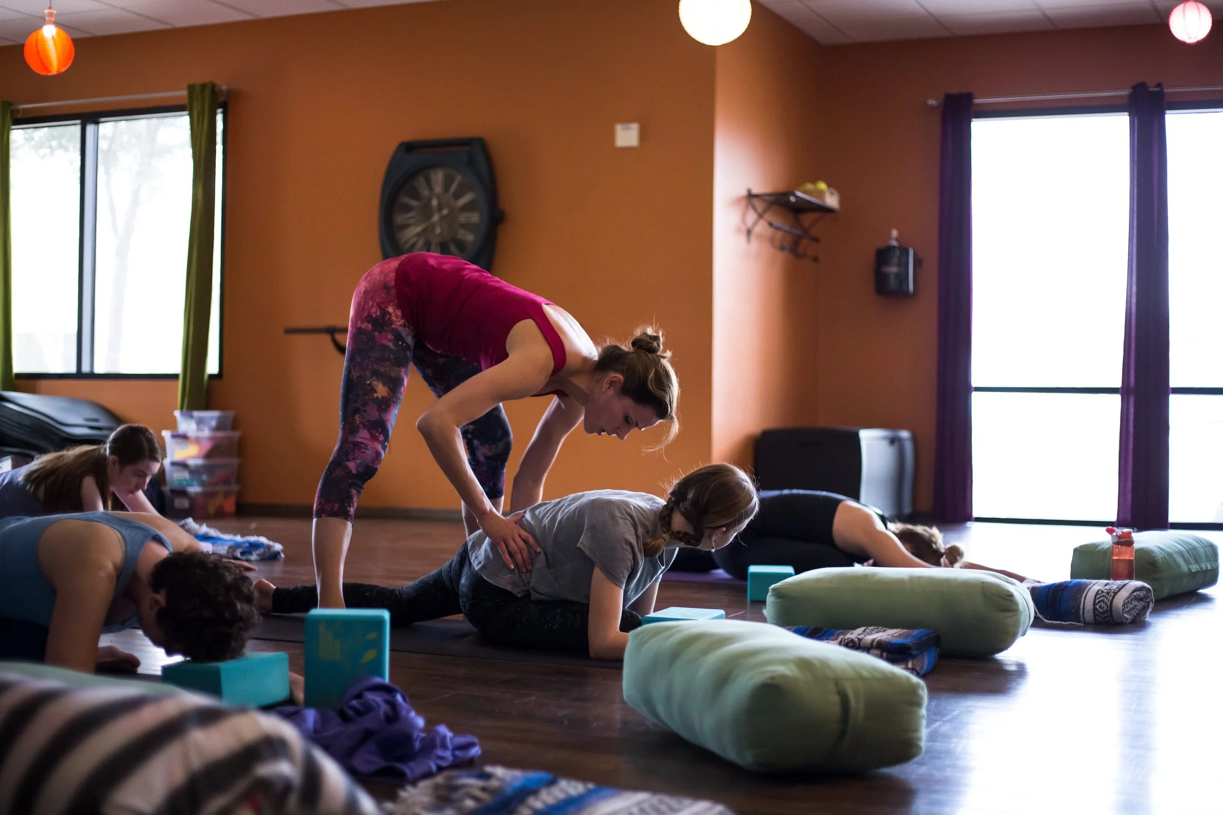 A yoga instructor assisting a mixed class with a variation of people with yoga poses in a studio with large windows and colorful decor.