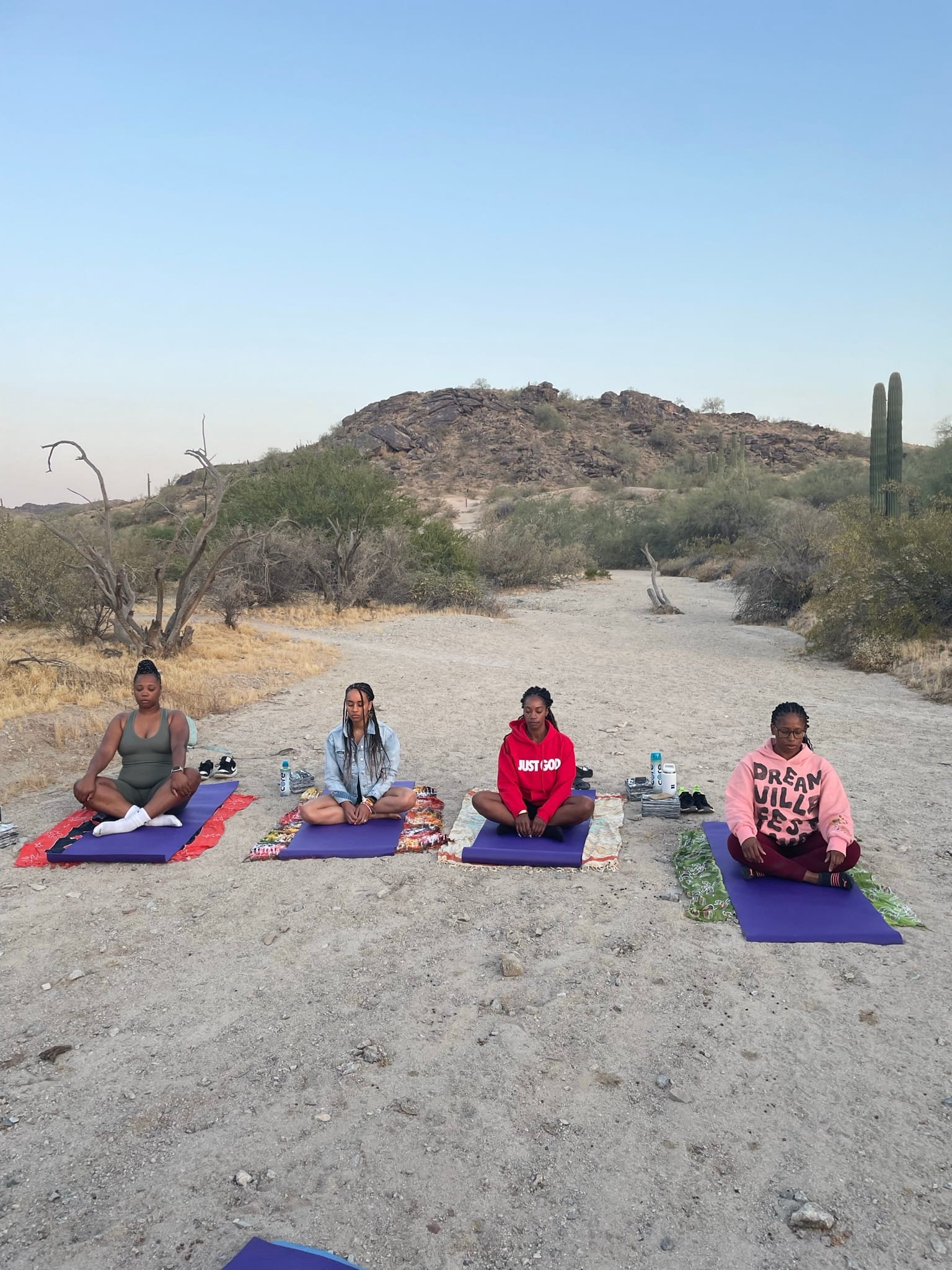 Four women practicing yoga in a desert landscape with cacti and rocky hills in the background during the early morning at sunrise.