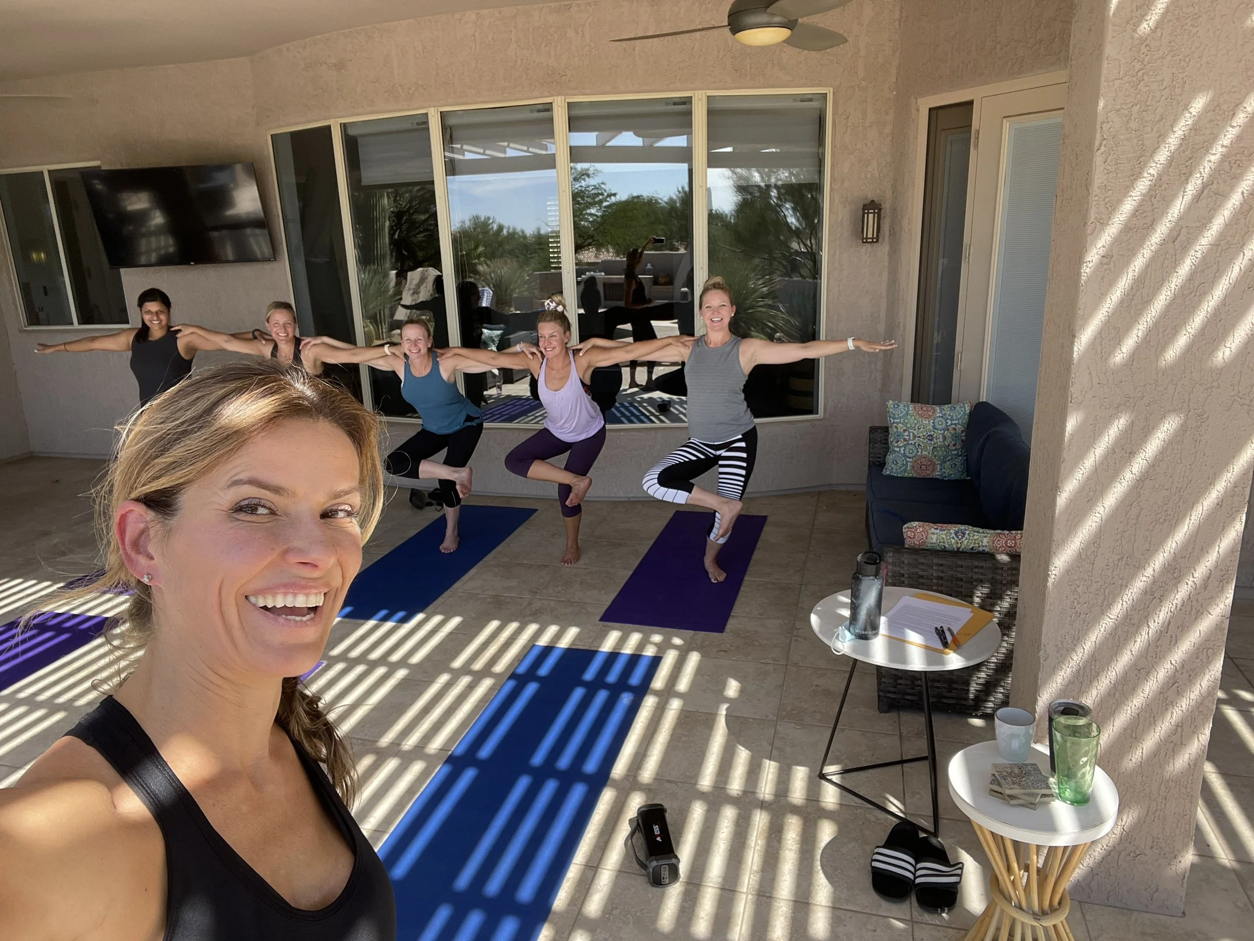 A woman takes a selfie during a yoga session with four women practicing yoga in the background on a covered outdoor patio, for a bachelorette party in Scottsdale, Arizona.