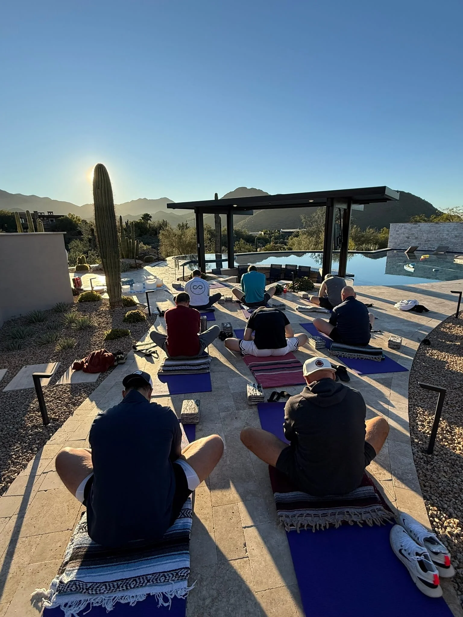 Men participating in an outdoor yoga session by a pool with mountains in the background during sunset for a yoga retreat.