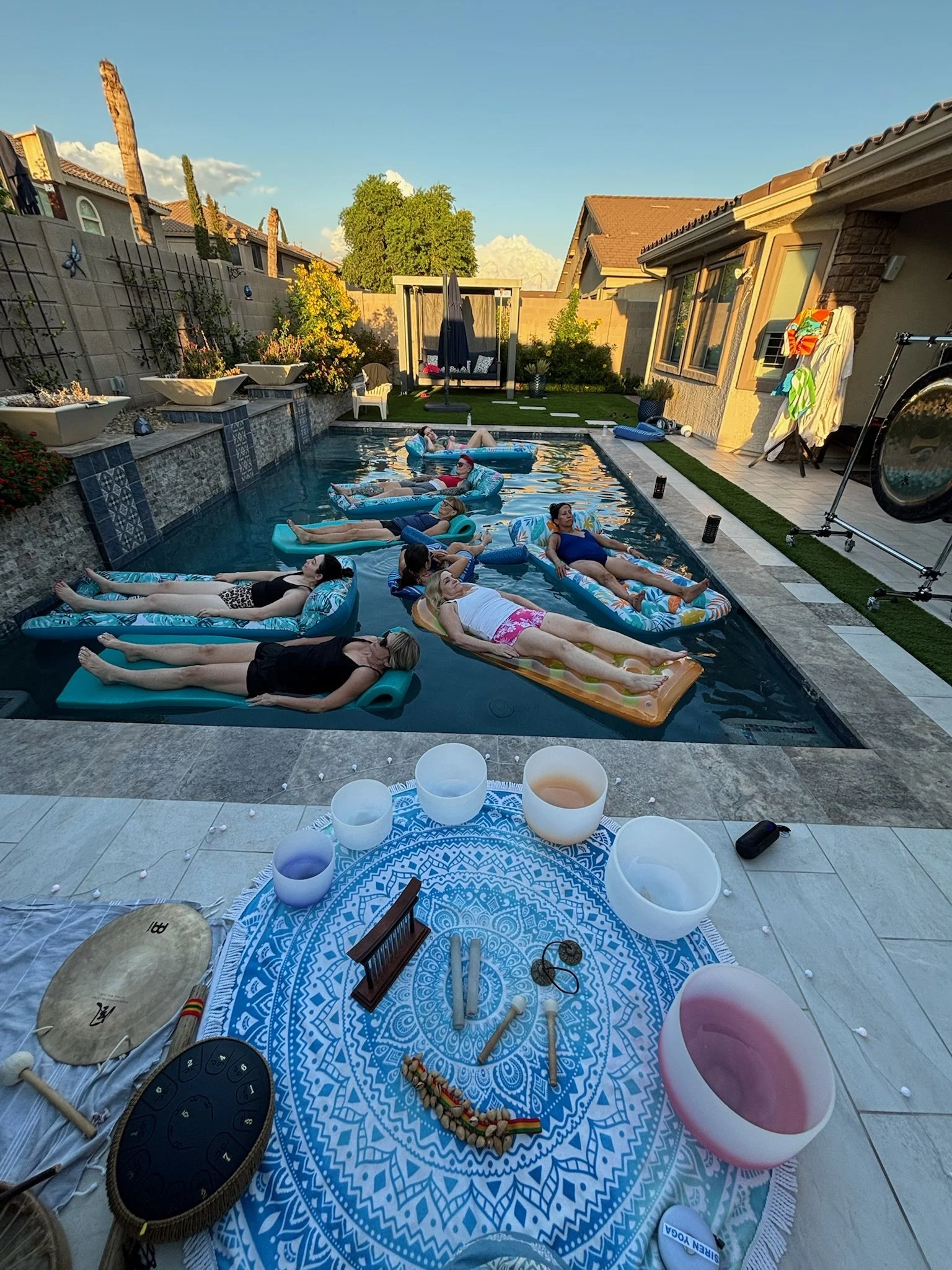 People relaxing on inflatable pool floats in a backyard swimming pool during a sunny day, with a decorated table and various musical instruments nearby.