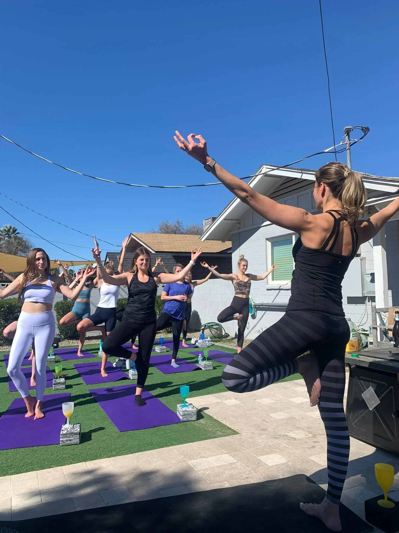 A group of women practicing yoga outdoors on purple mats under a clear blue sky, with an instructor leading the session.