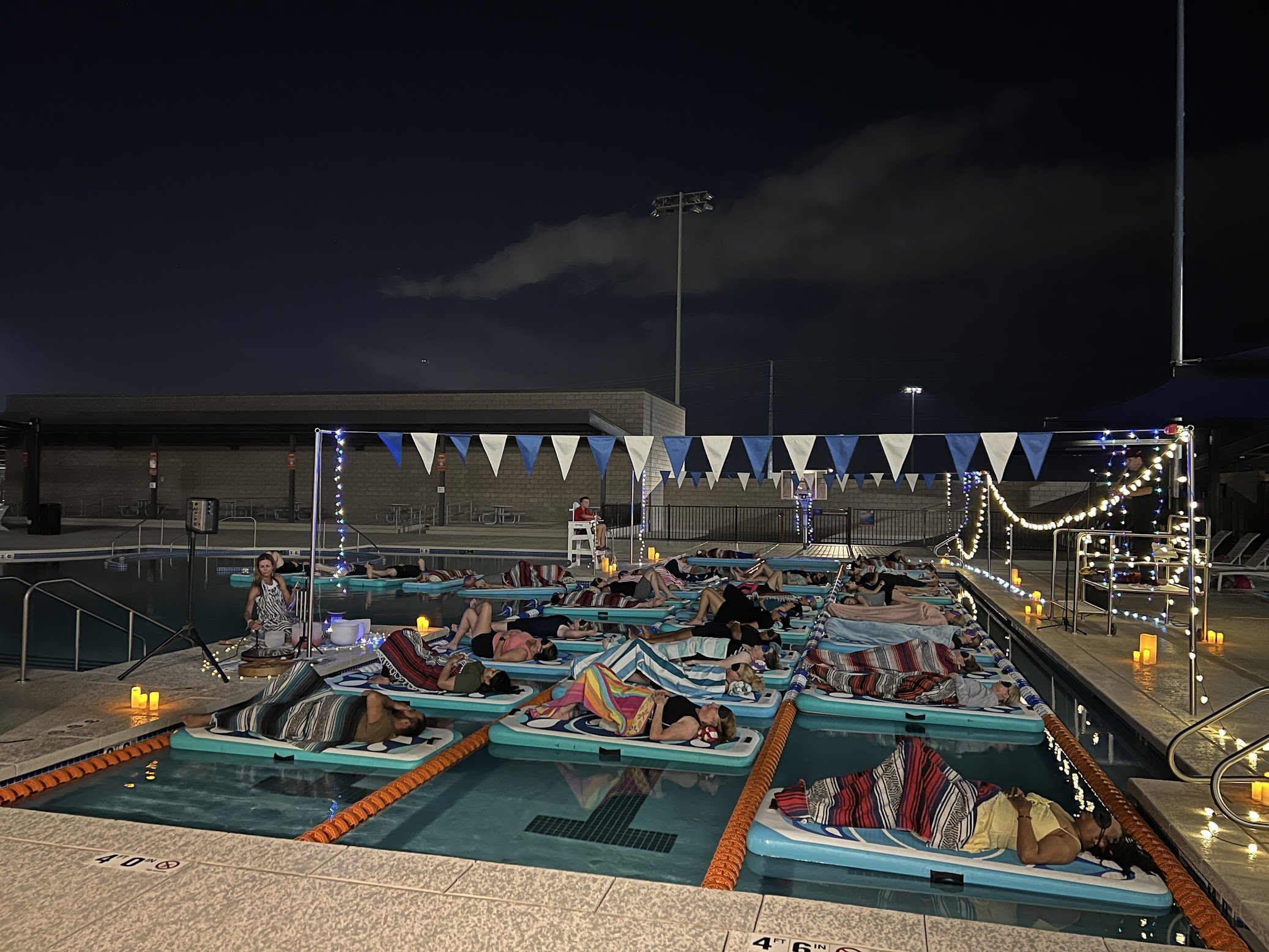 People lounging on floating beds in an outdoor swimming pool at night, decorated with string lights and pennant banners, with a lifeguard on a chair and candles around the pool area.