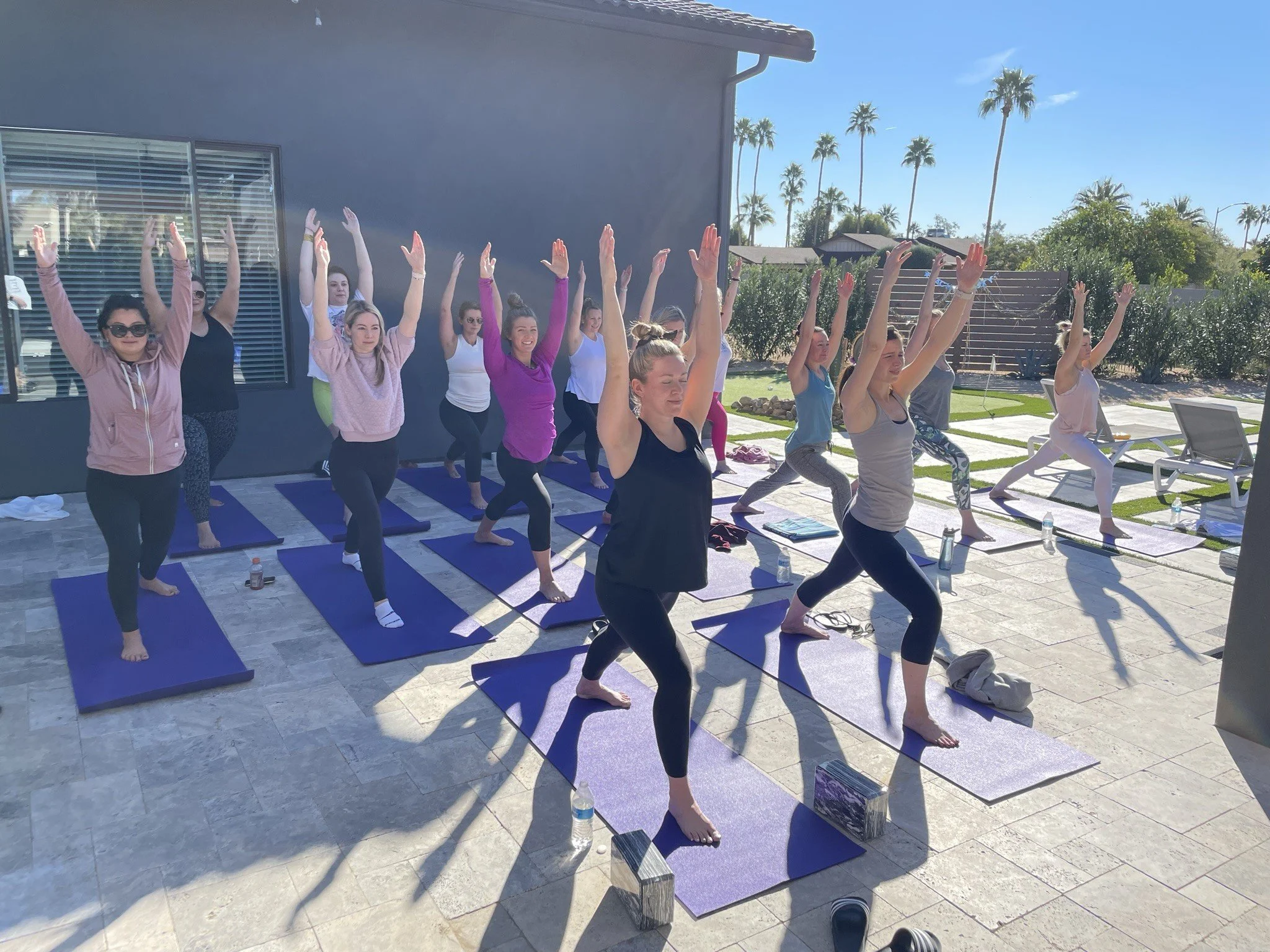 A diverse group of women practicing yoga outdoors on yoga mats. They are in a downward dog pose with their arms raised and looks of concentration and relaxation. The setting appears sunny and warm in an Airbnb in Scottsdale in Arizona.