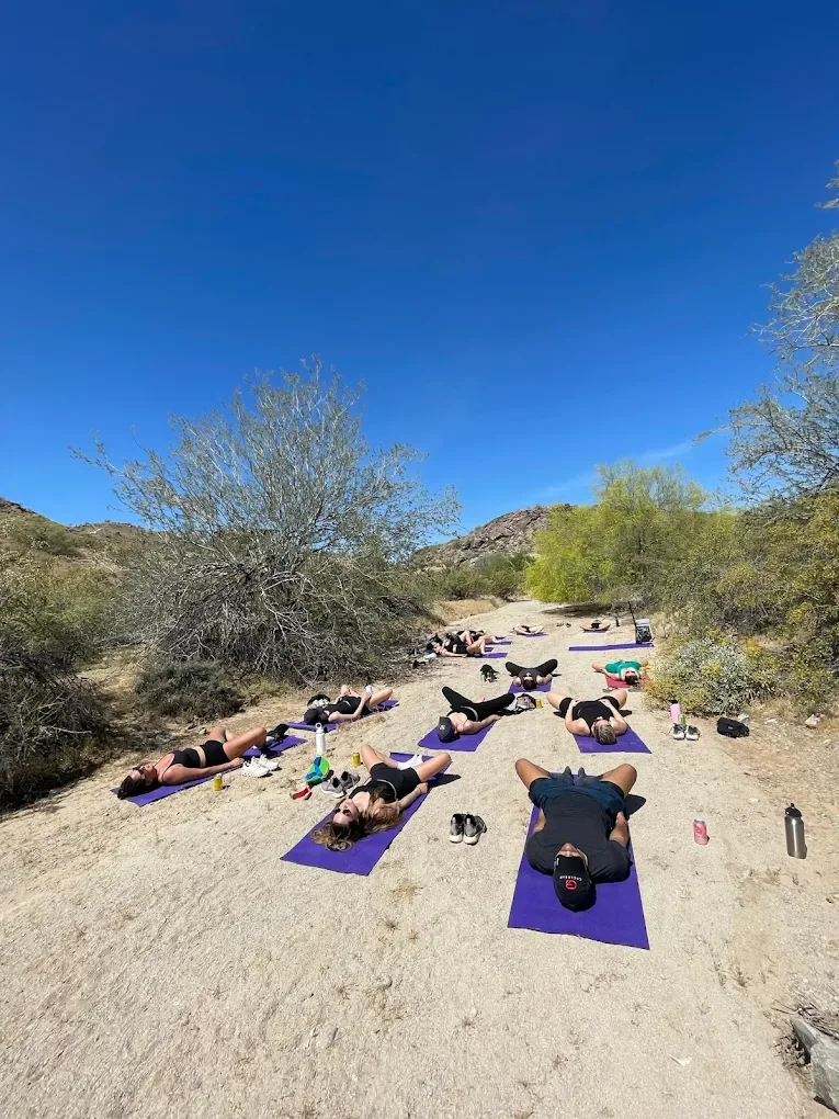 People practicing yoga on purple mats outdoors in a desert landscape with sparse trees and mountains under a clear blue sky after a Hike and Yoga session.
