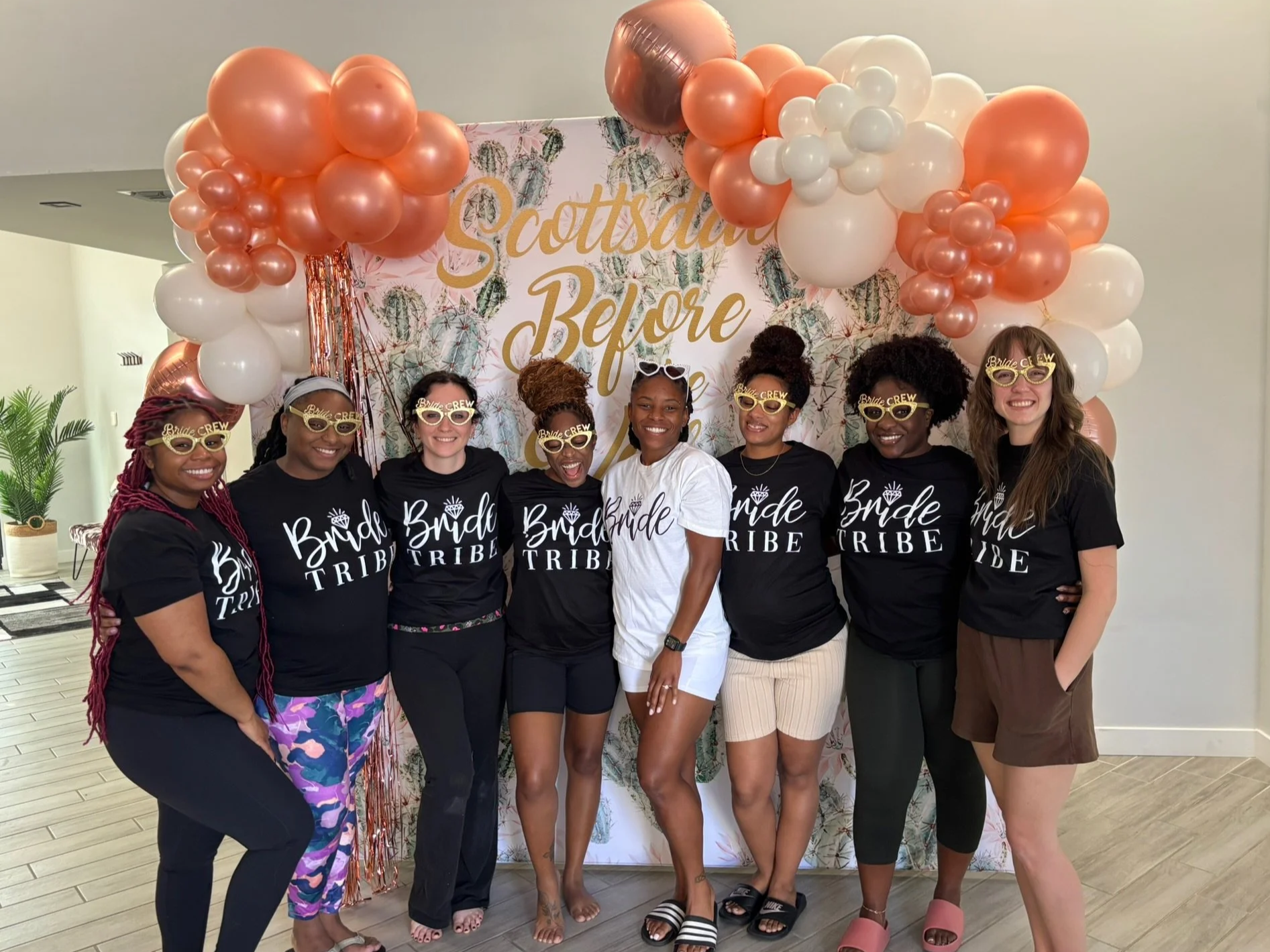 A group of eight women celebrating wearing matching black T-shirts that read 'Bride Tribe' and fun yellow glasses. They are standing in front of a decorated backdrop with photo balloons and a sign that says 'Scottsdale Before the Veil" .