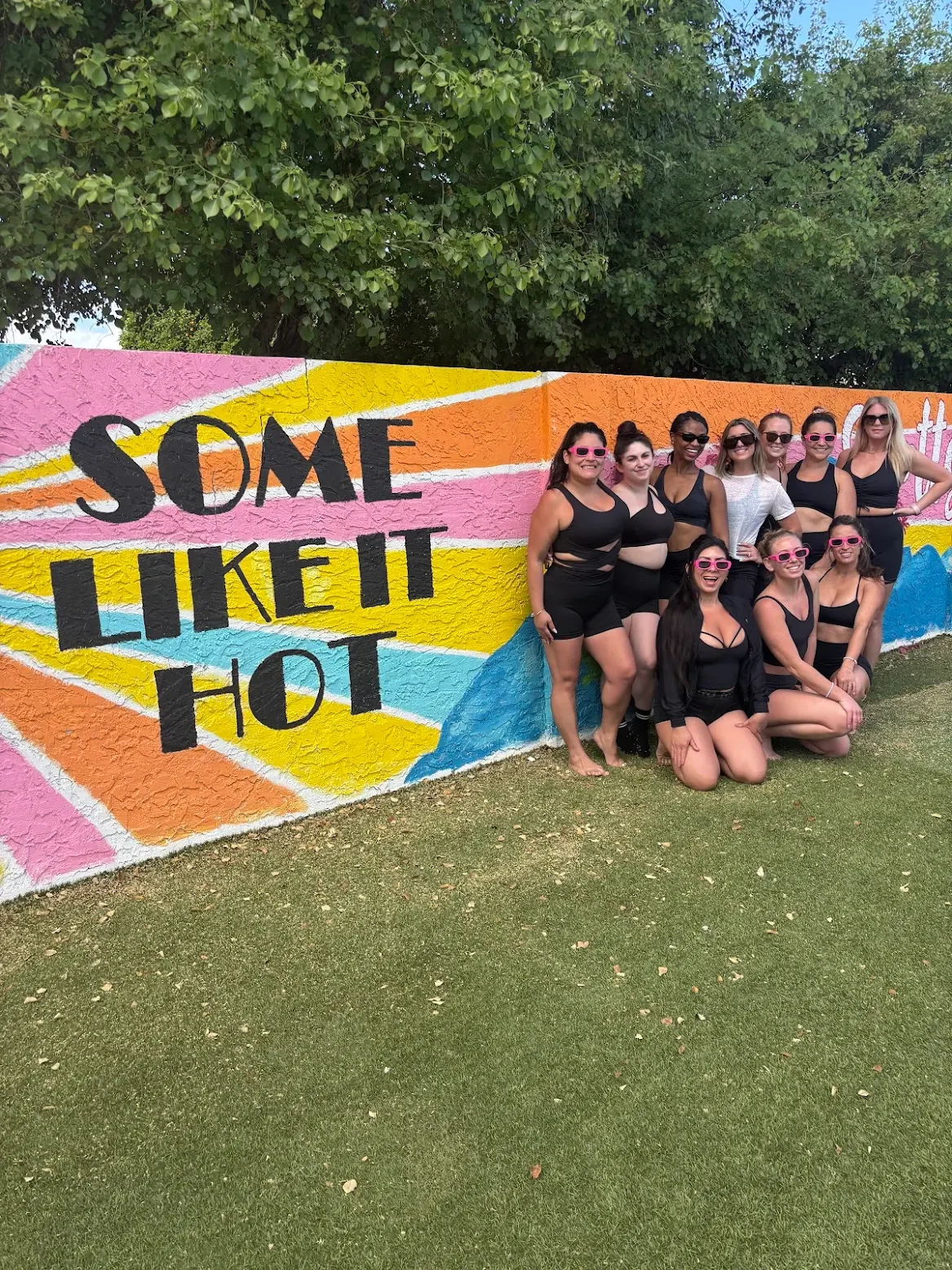 Group of women in black outfits and pink sunglasses posing prior to an outdoors Bachelorette Yoga session in front of a colorful mural with the phrase 'Some Like It Hot'