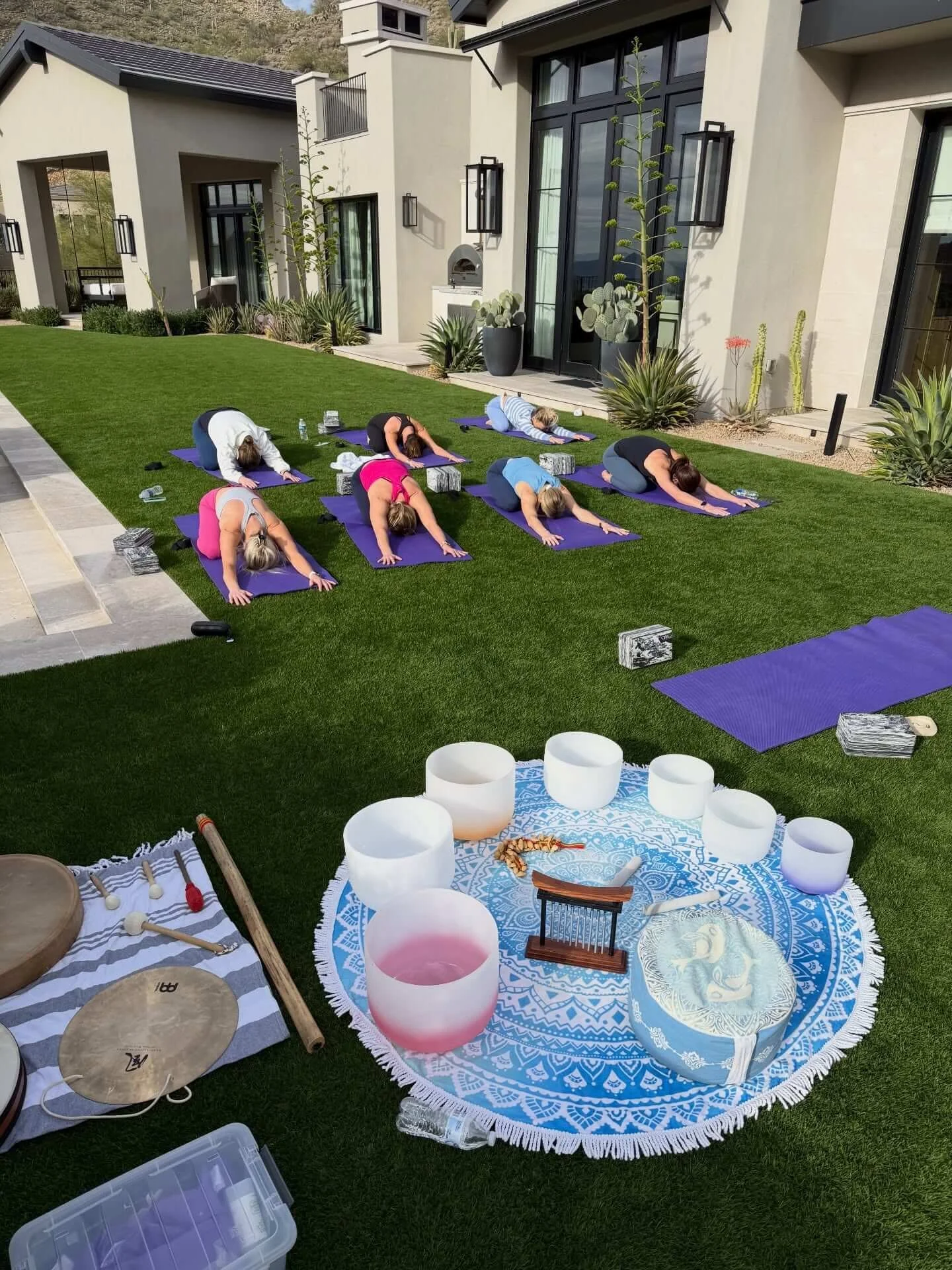 Group of women practicing yoga on outdoor purple mats in a backyard, seated in a cross-legged meditation pose with eyes closed, sunlight, and modern patio furniture celebrating a Bachelorette.