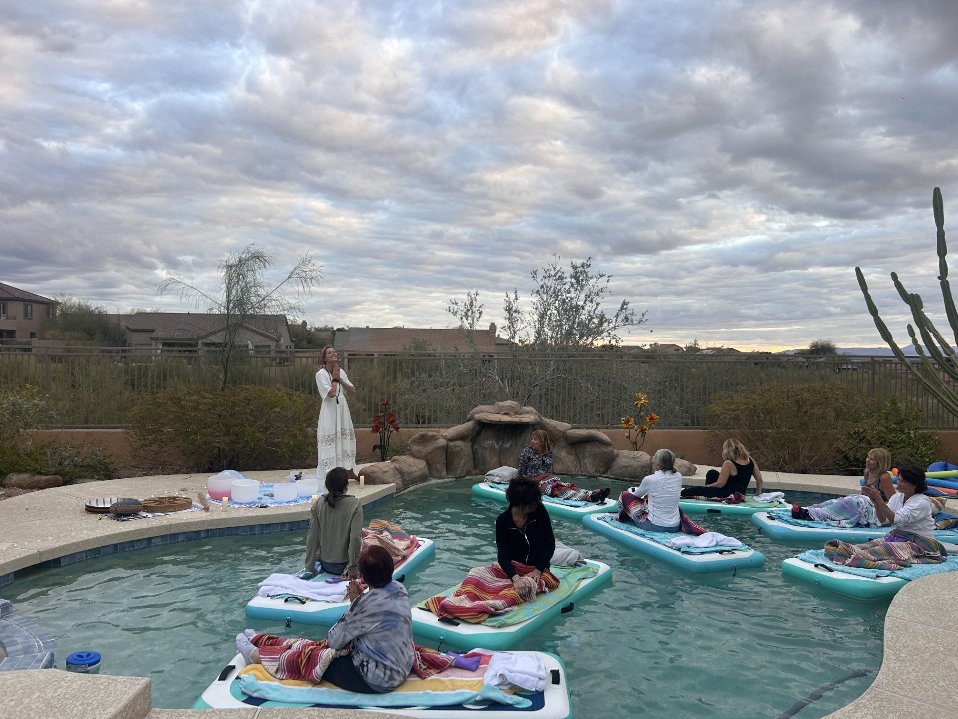 A group of people participating in a meditation or yoga session on paddleboards in a swimming pool outdoors, with a caregiver playing singing bowls.