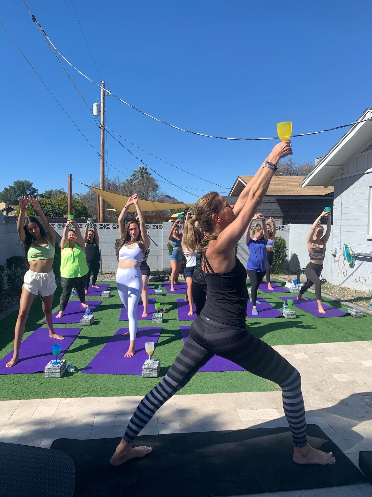 Group of women practicing yoga outdoors on purple mats on a sunny day. They are stretching and holding glasses, with Vanessa Siren from Siren Yoga leading at the front. All are dressed in athletic wear for a Bachelorette Yoga session.