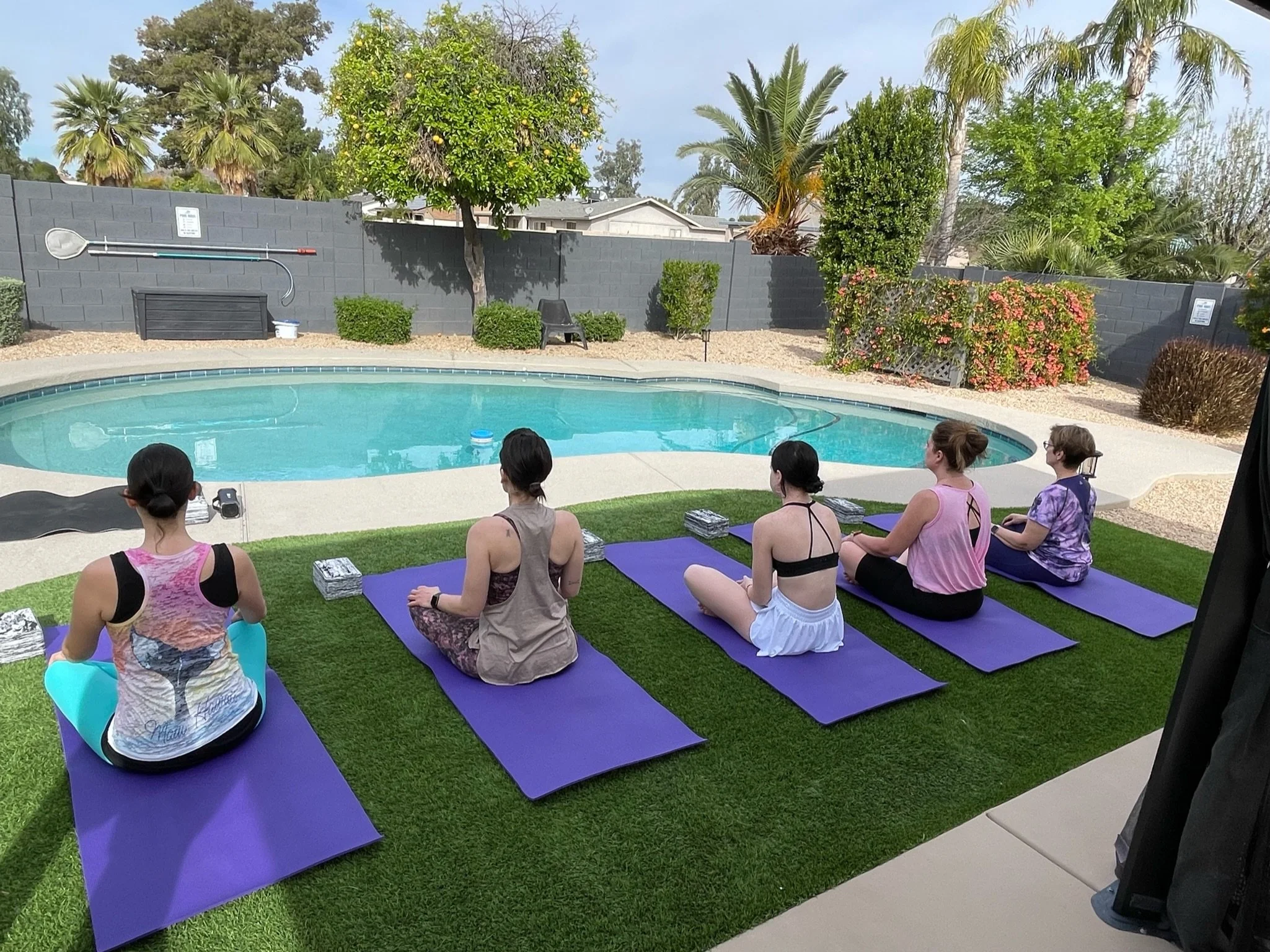 Five women in bachelorette yoga posing on purple mats practicing outside near a swimming pool, with trees and plants in the background at their Airbnb.
