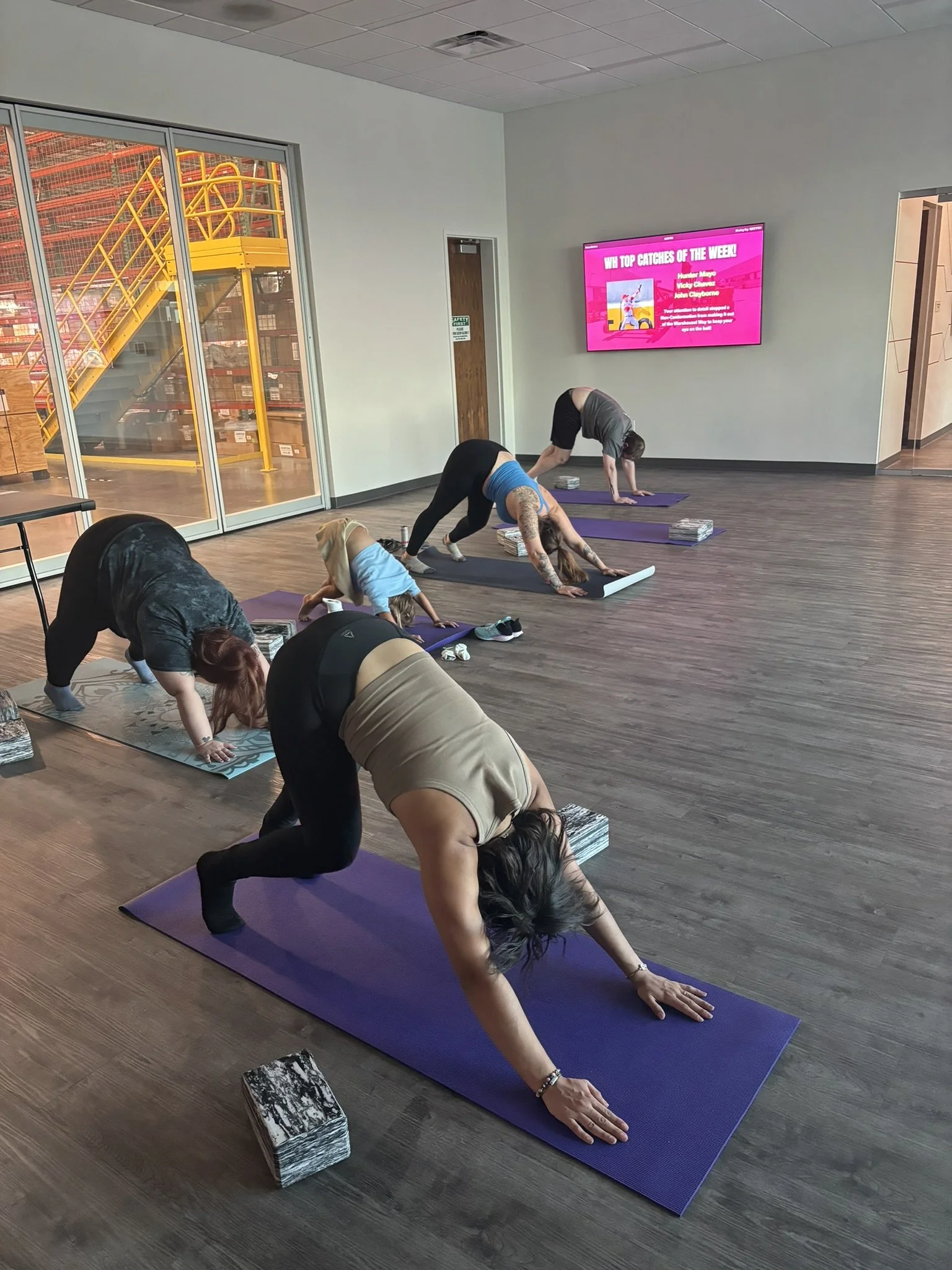 People participating in a yoga class practicing downward dog pose on mats in a corporate setting next to a warehouse window of a factory.