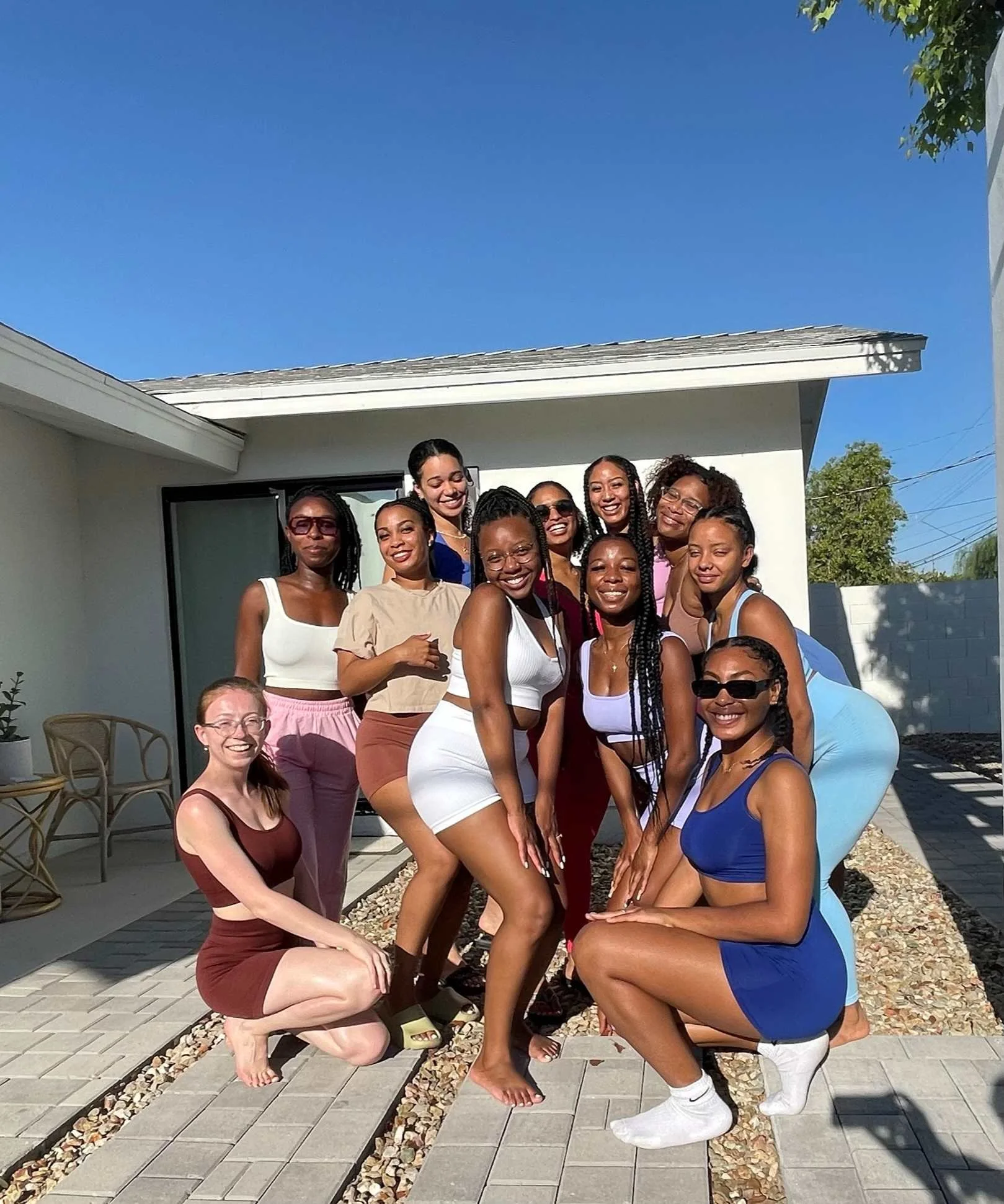 Group of women posing outdoors in front of a white house on sunny day, smiling in yoga clothes ready for their outdoor practice