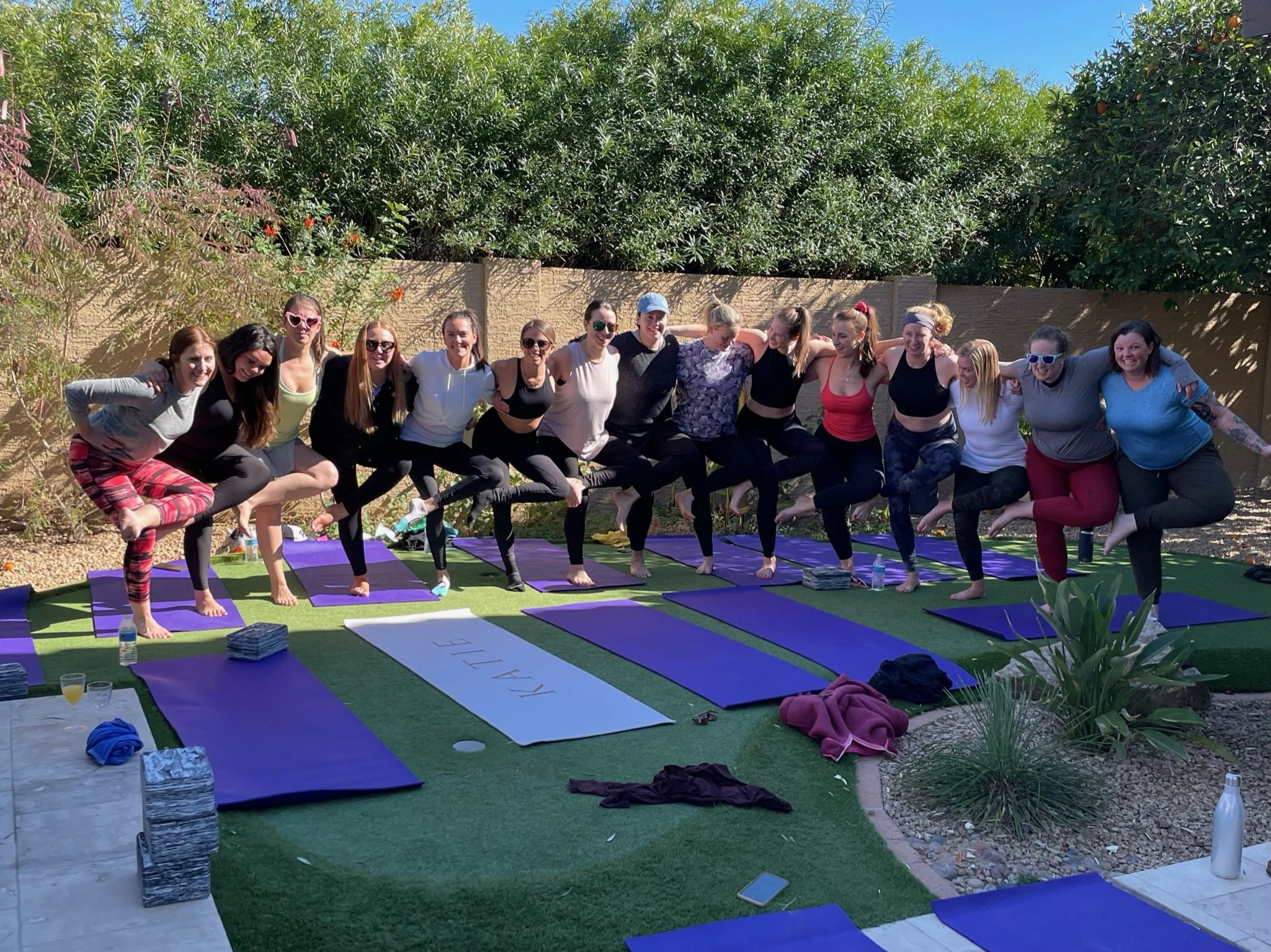 A group of women practicing bachelorette yoga outdoors on purple mats, standing in a line with their arms around each other's shoulders, in a garden setting with trees and a wooden fence in the background.