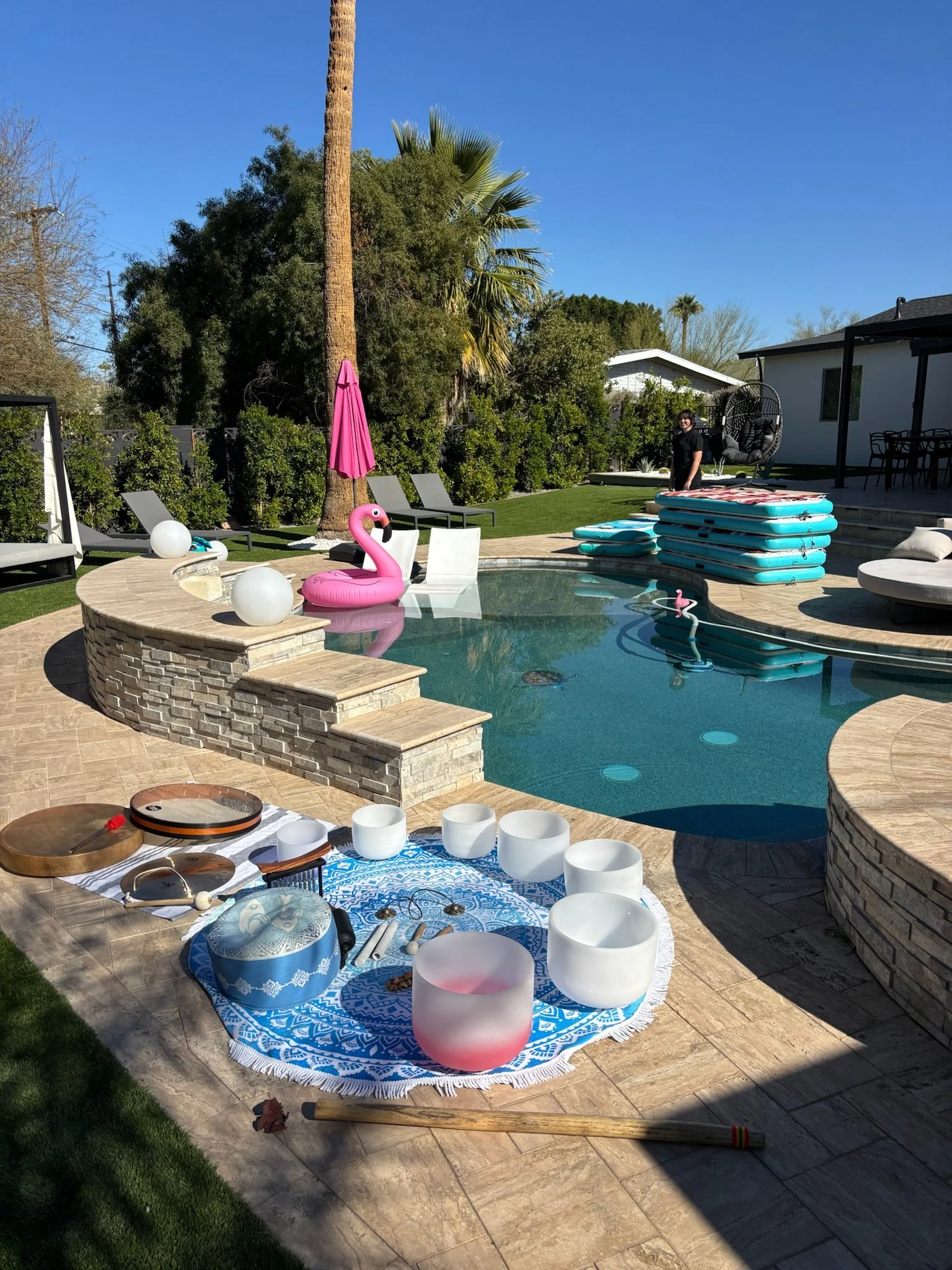A backyard swimming pool with a pink flamingo float, poolside lounge chairs, and inflatable pool floats. There are musical instruments, bowls used as sound bowls, and a person standing near the pool. The scene is sunny with a blue sky and green trees
