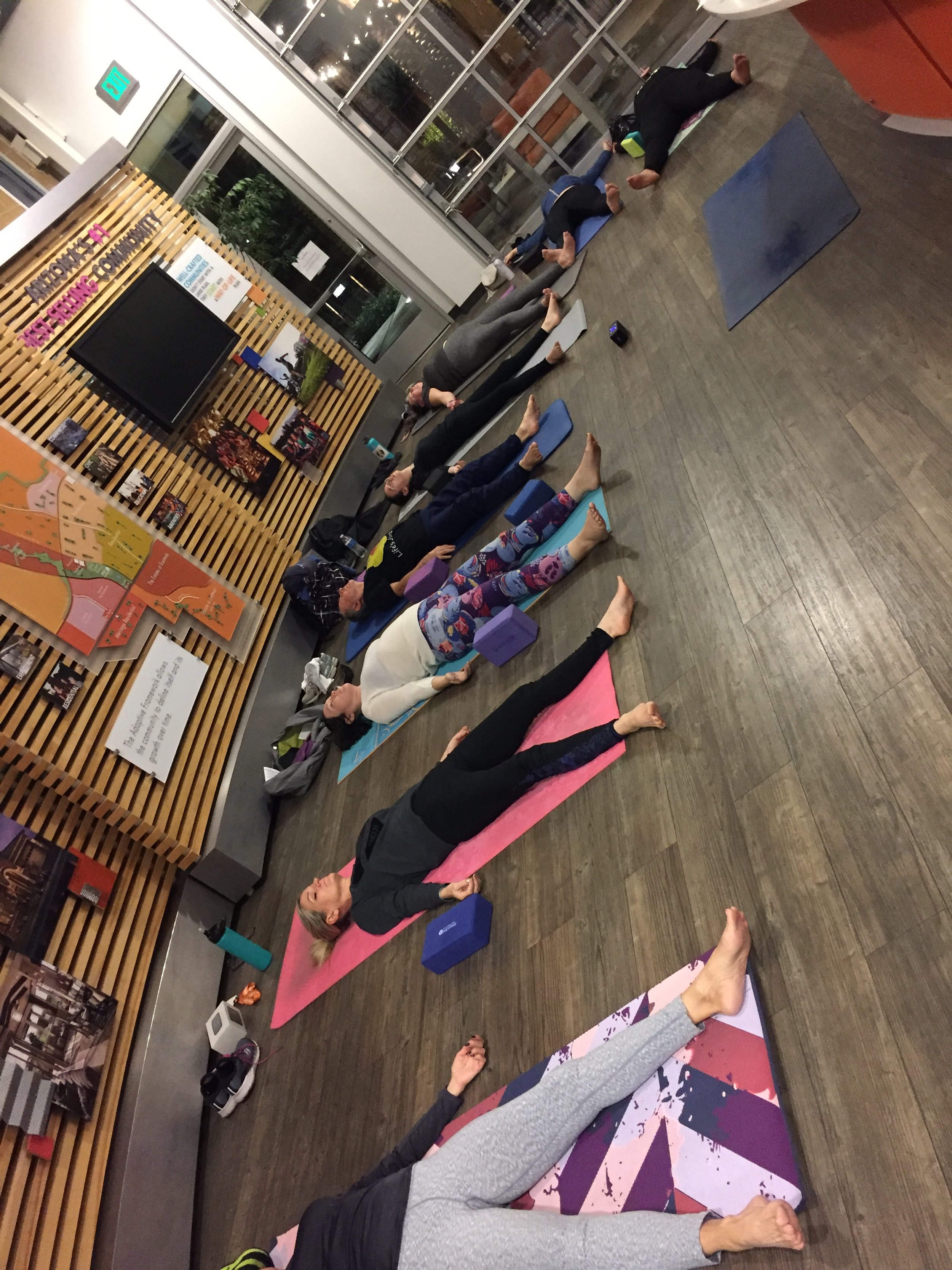 People participating in a yoga class indoors, lying on yoga mats with yoga blocks, in front of a wooden slat wall with a display board and artwork for a corporate private session.
